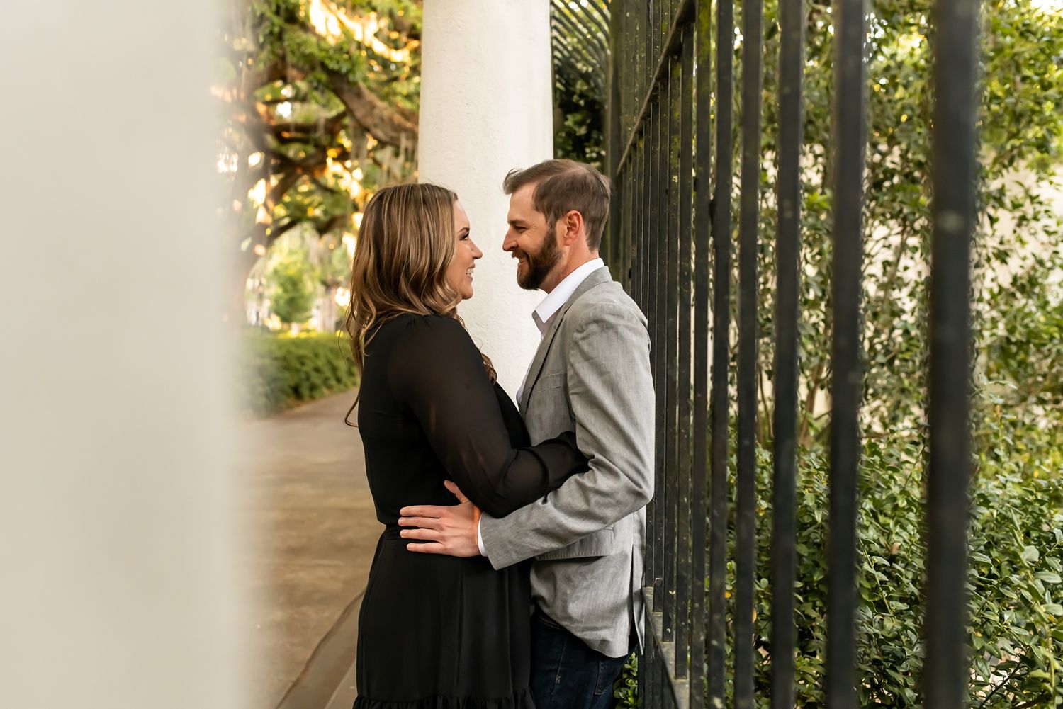 A couple shares an intimate moment against a white pillar and decorative railing surrounded by greenery.
