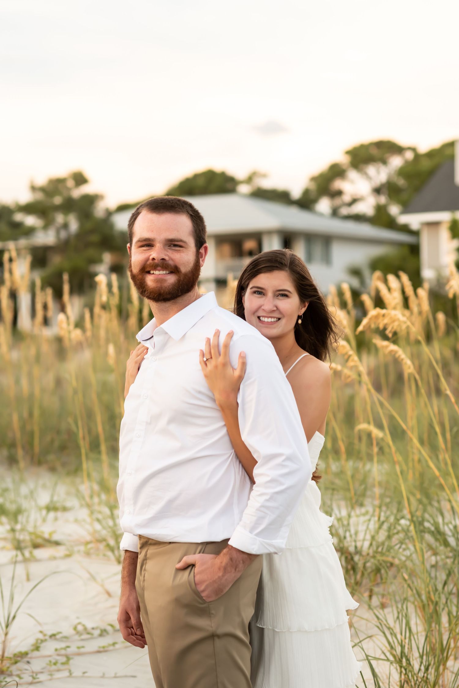 Happy couple in casual wedding attire pose together on a beach with sea grass during golden hour.