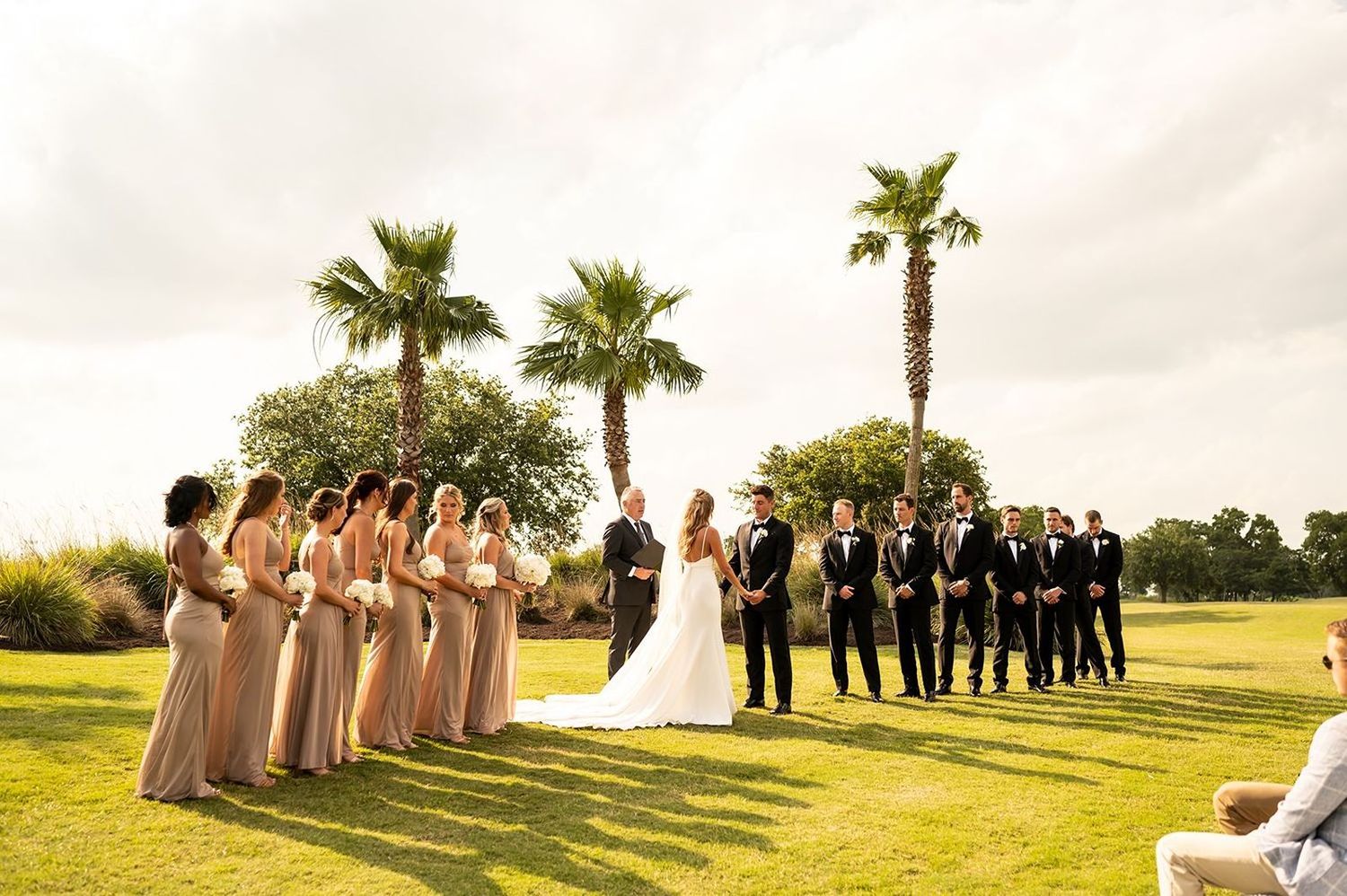 Outdoor wedding ceremony on a golf course with palm trees and bridesmaids in blush dresses and groomsmen in dark suits.
