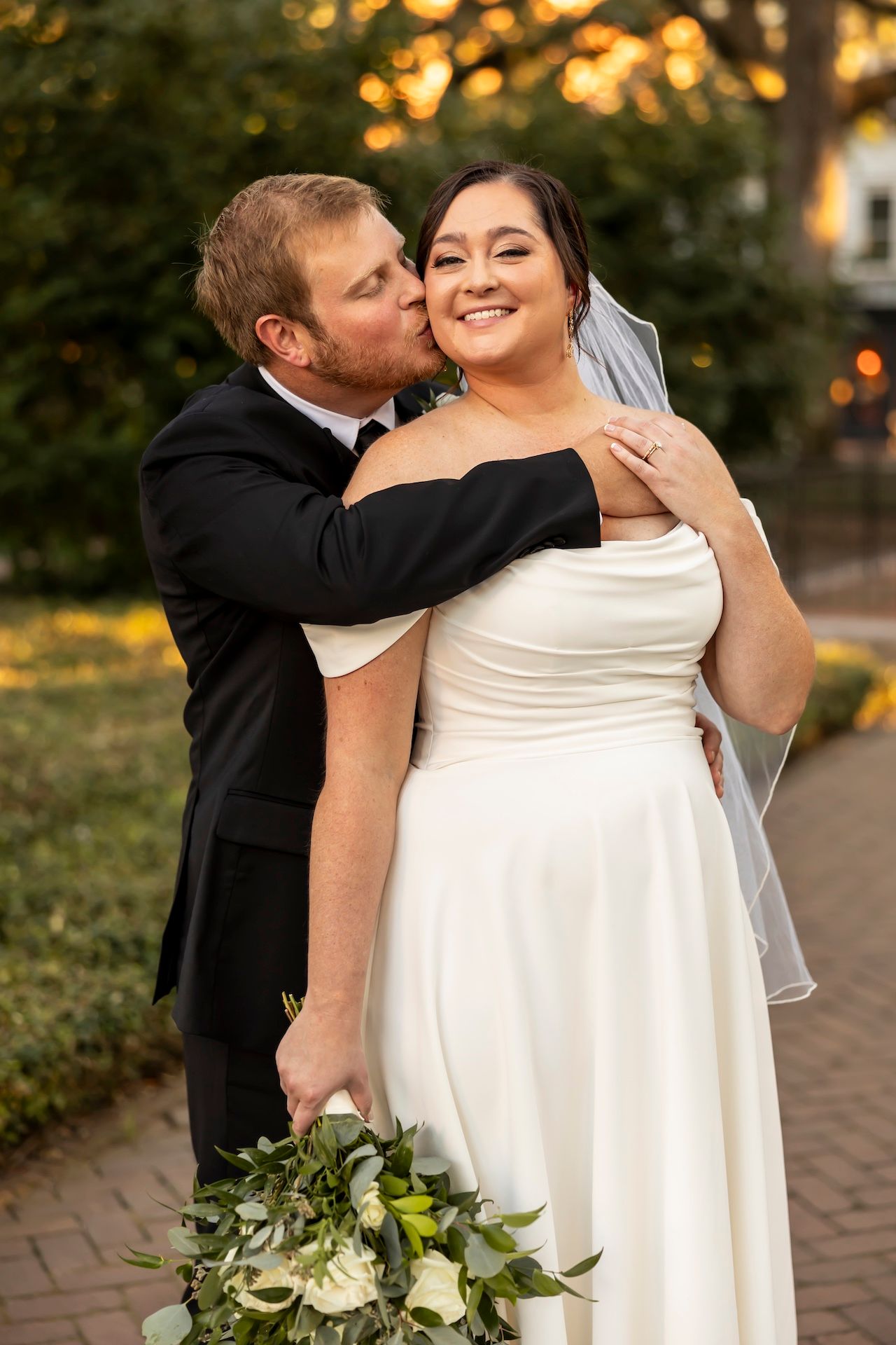 Newlywed couple sharing a tender moment outdoors with bride holding white and green bouquet at sunset.