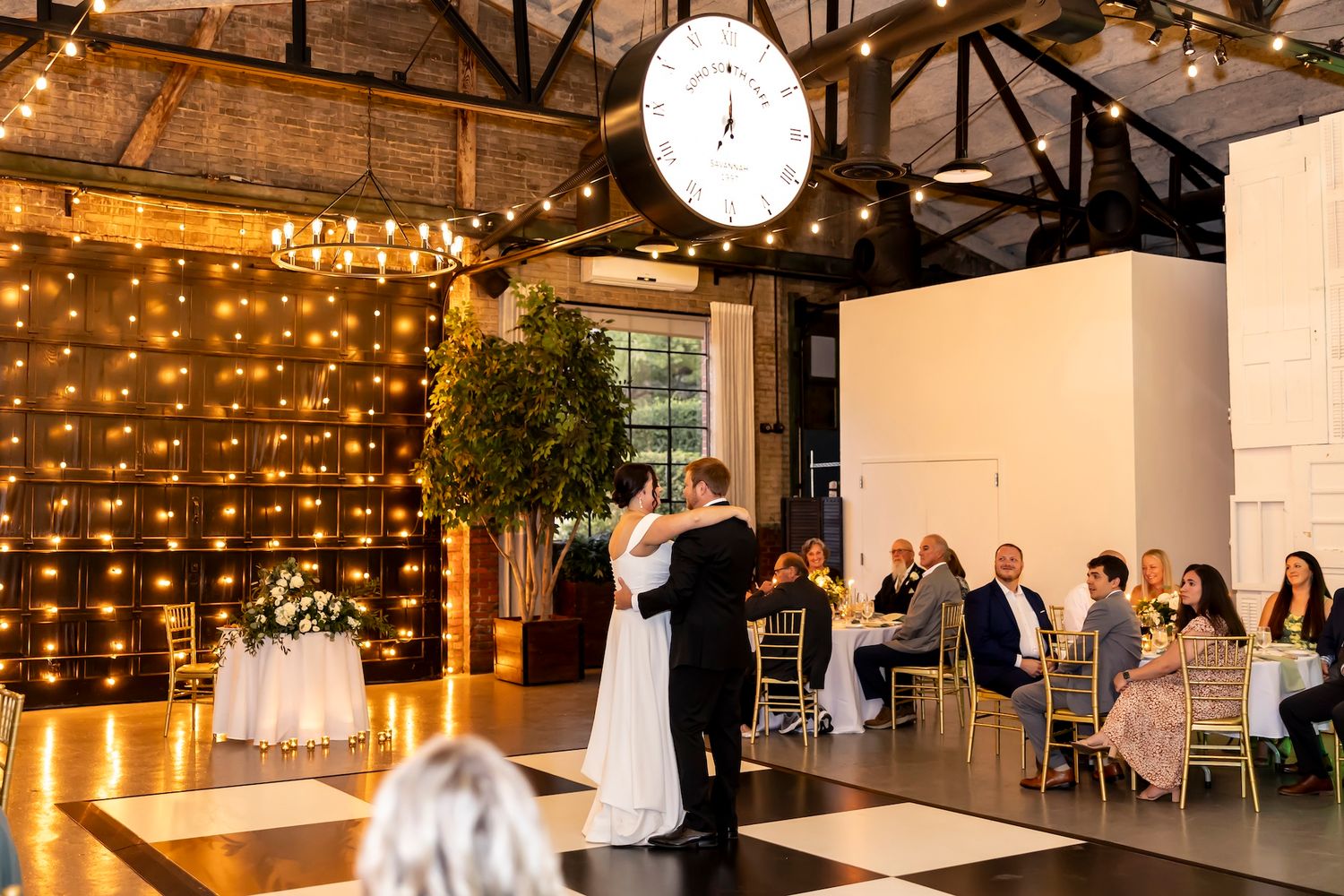 Wedding couple shares their first dance in an elegantly lit industrial venue with guests seated at dinner tables.
