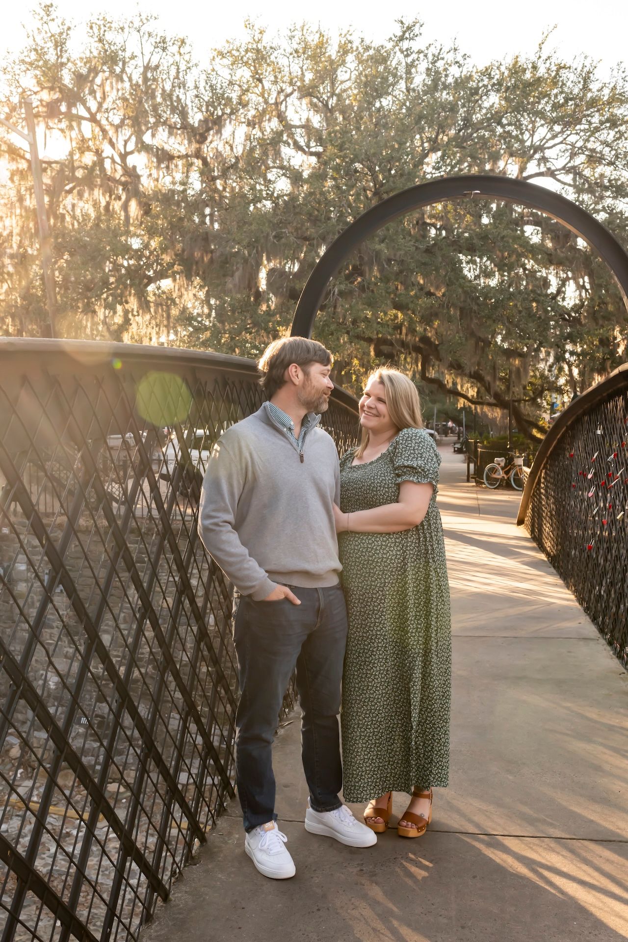 A couple shares a romantic moment on a wrought iron bridge at sunset with Spanish moss draped trees in the background.