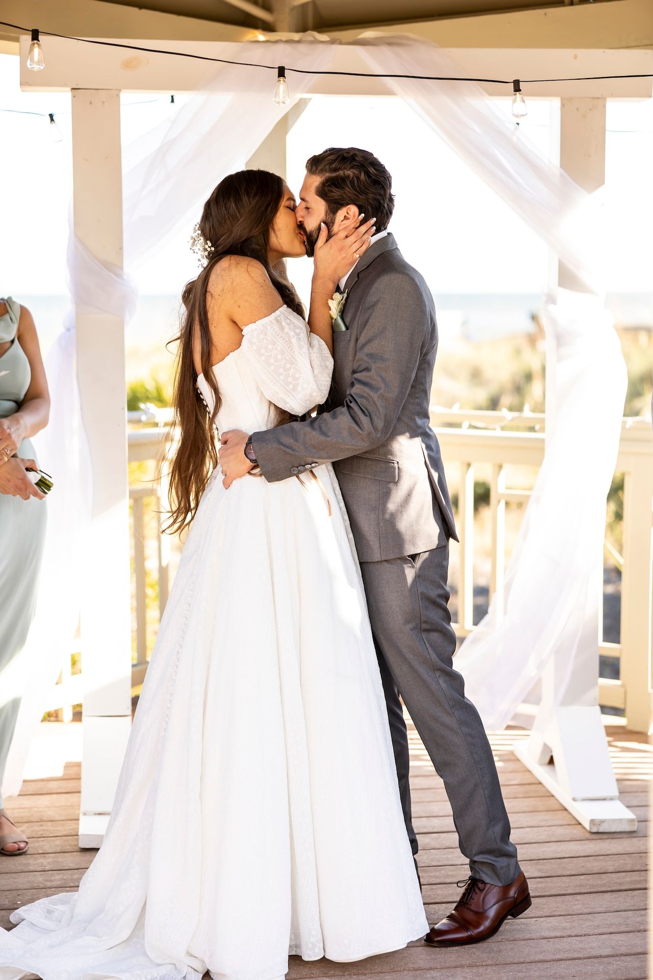 A bride and groom share a romantic kiss on a covered deck during their wedding day ceremony in soft sunlight.
