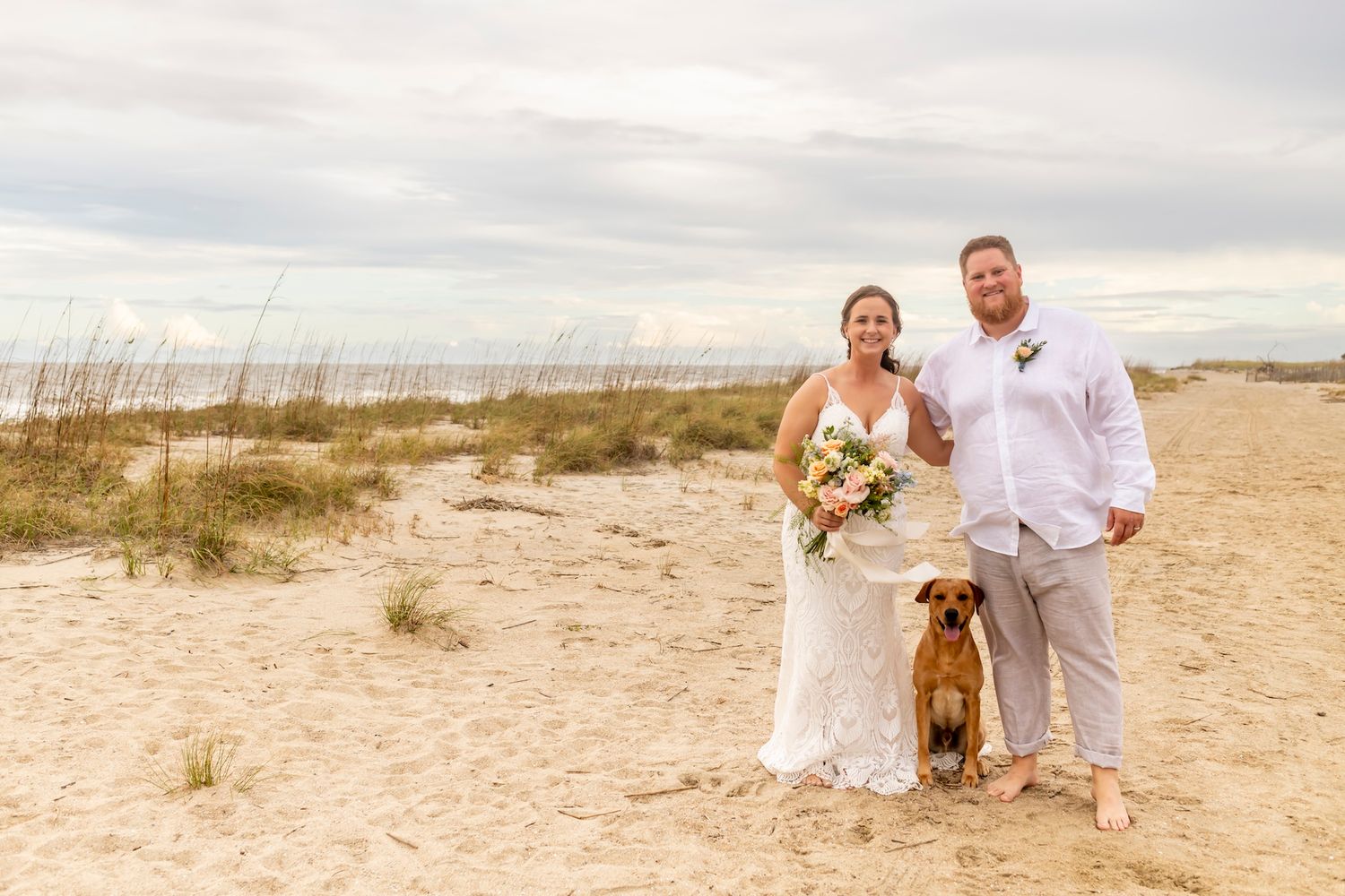 A couple celebrates their beach wedding with their brown dog on a sandy dune with sea grass in the background.