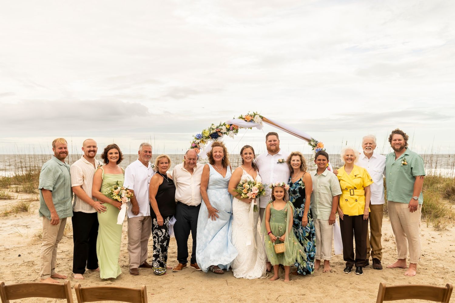 Wedding party gathered on wooden boardwalk against beachfront backdrop with decorated arch and ocean view.