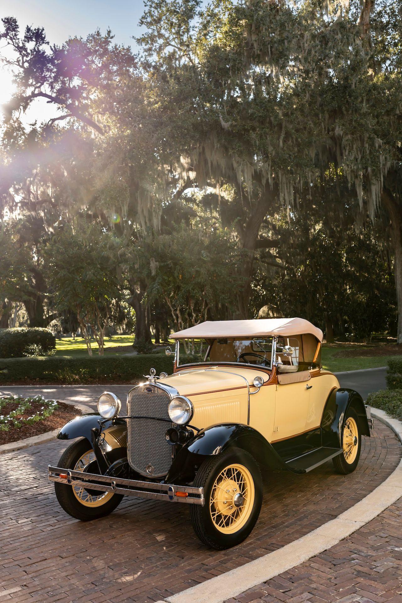 A vintage yellow Ford Model A convertible parked on a brick road under Spanish moss-draped trees in golden sunlight.