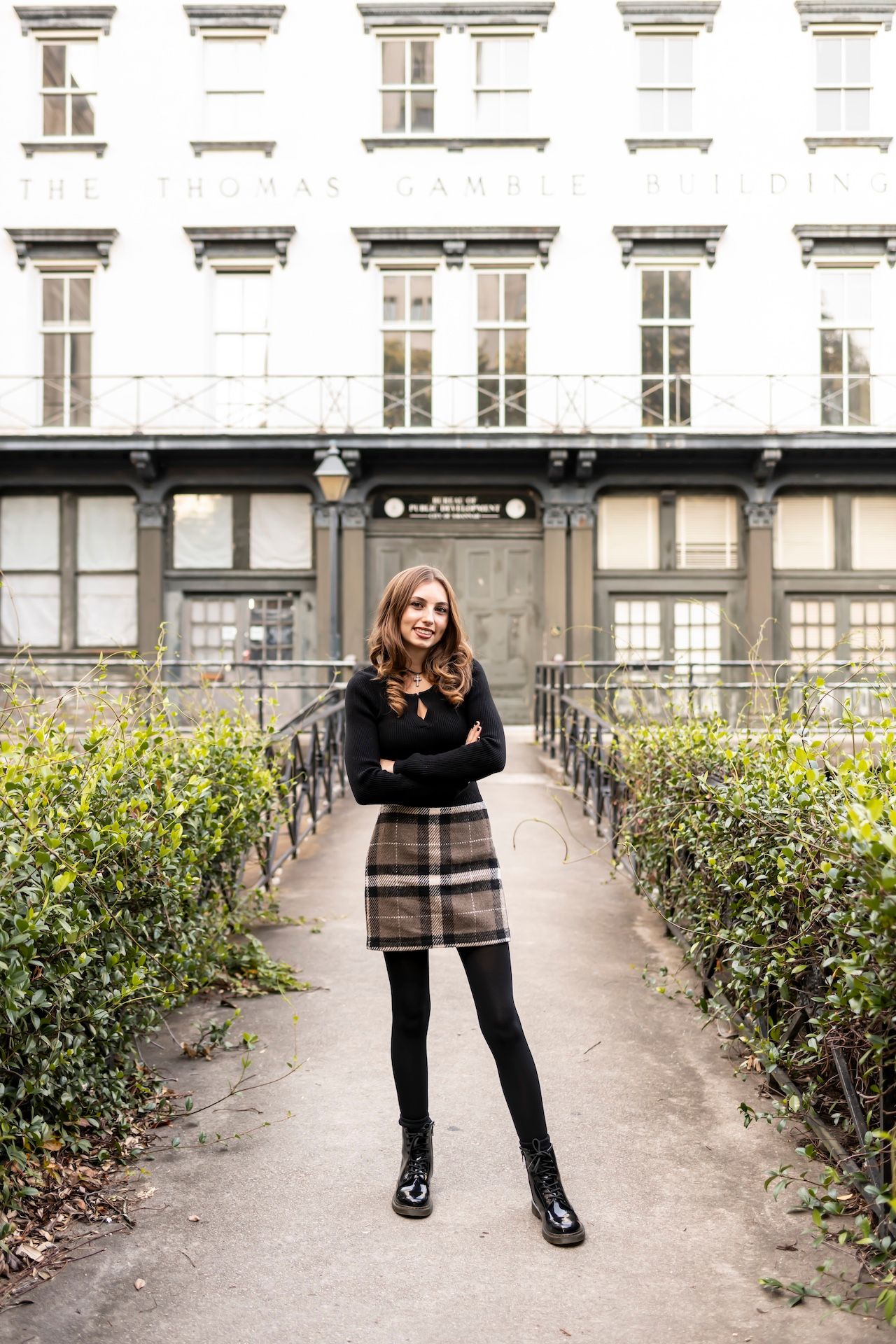 Person wearing black sweater and plaid skirt poses on garden path in front of historic building with elegant windows.
