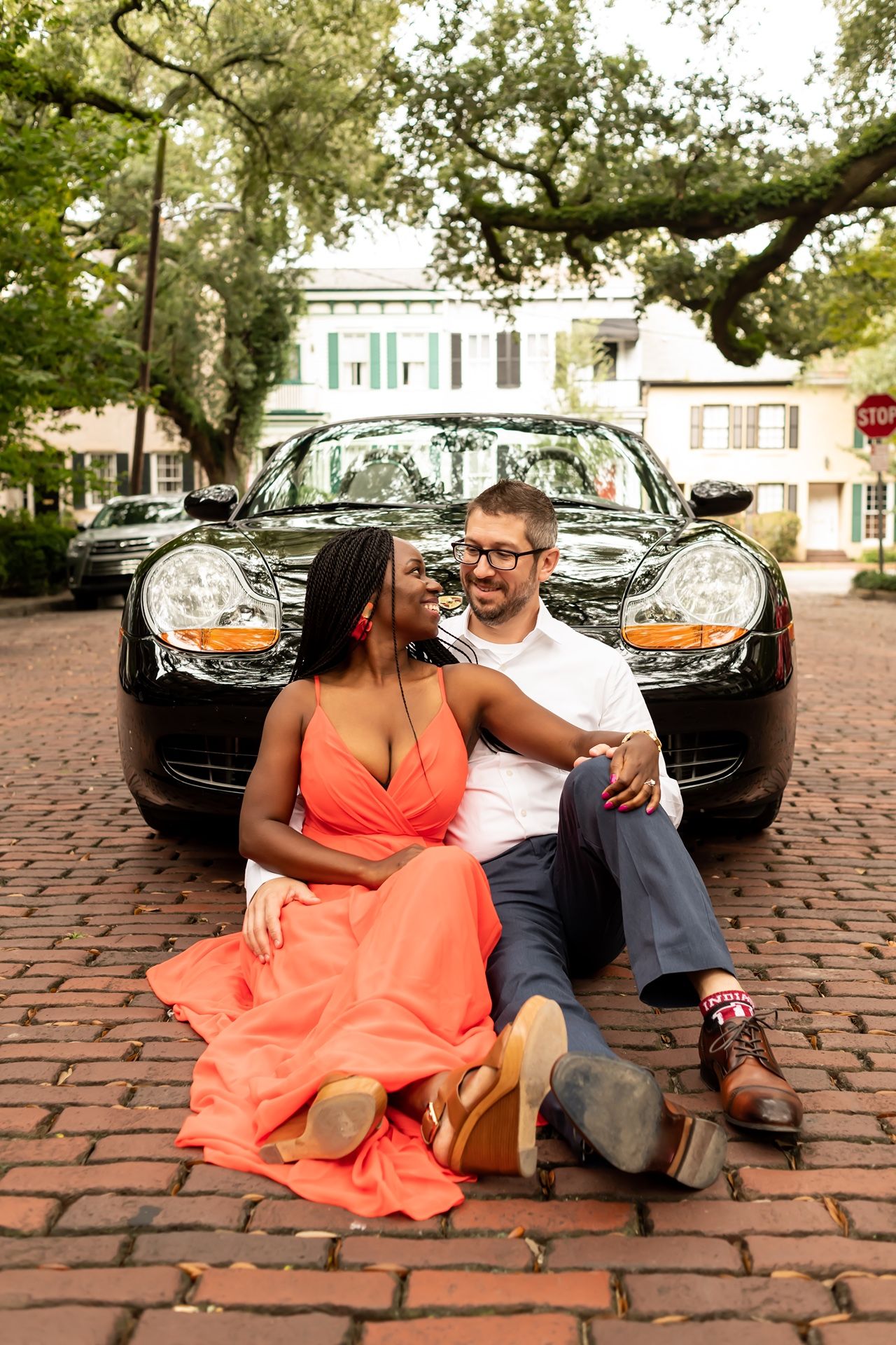 A romantic couple sits on cobblestone pavement next to a black car, with lush trees and historic buildings in the background.