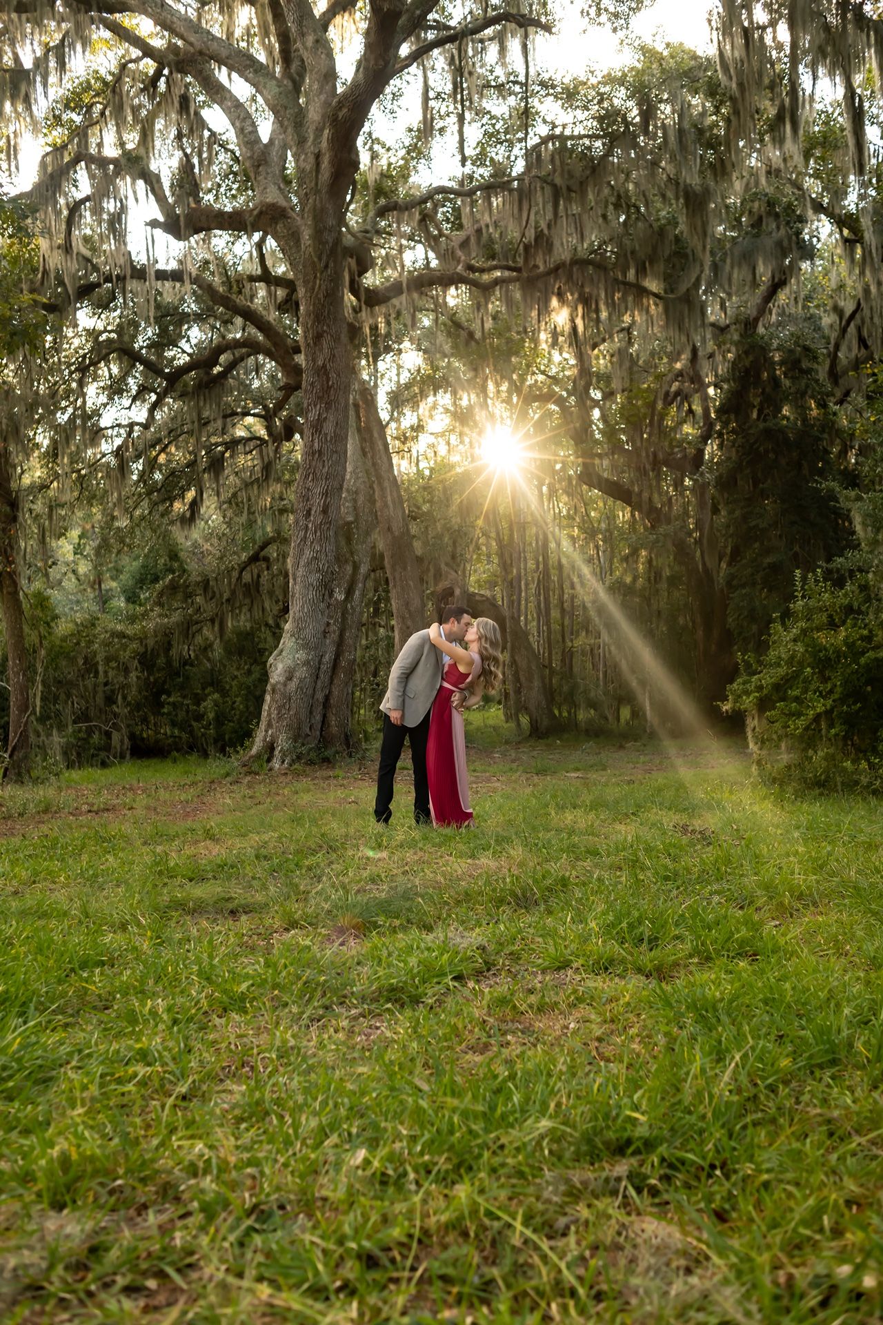Sunlight streams through Spanish moss-draped trees as a couple embraces in a dreamy outdoor setting.