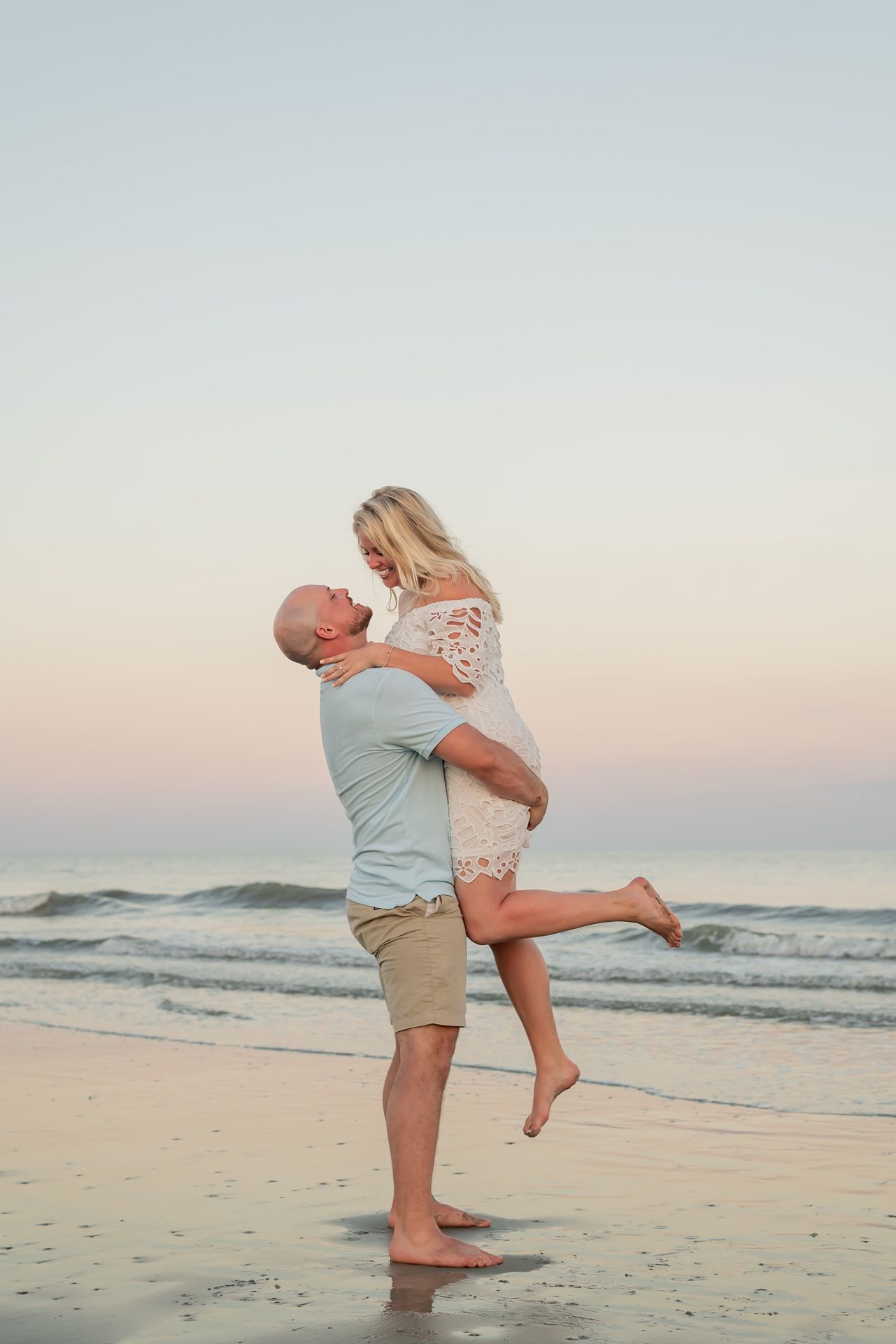 A couple embraces and dances together at sunset on a peaceful beach with rolling waves in the background.