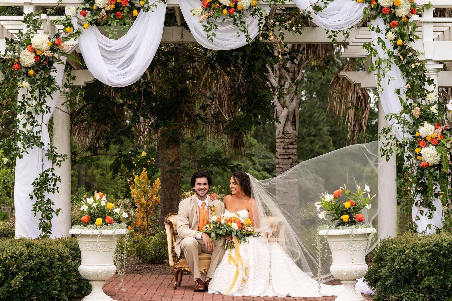 A romantic wedding ceremony under a white floral arch decorated with orange blooms and flowing white fabric.