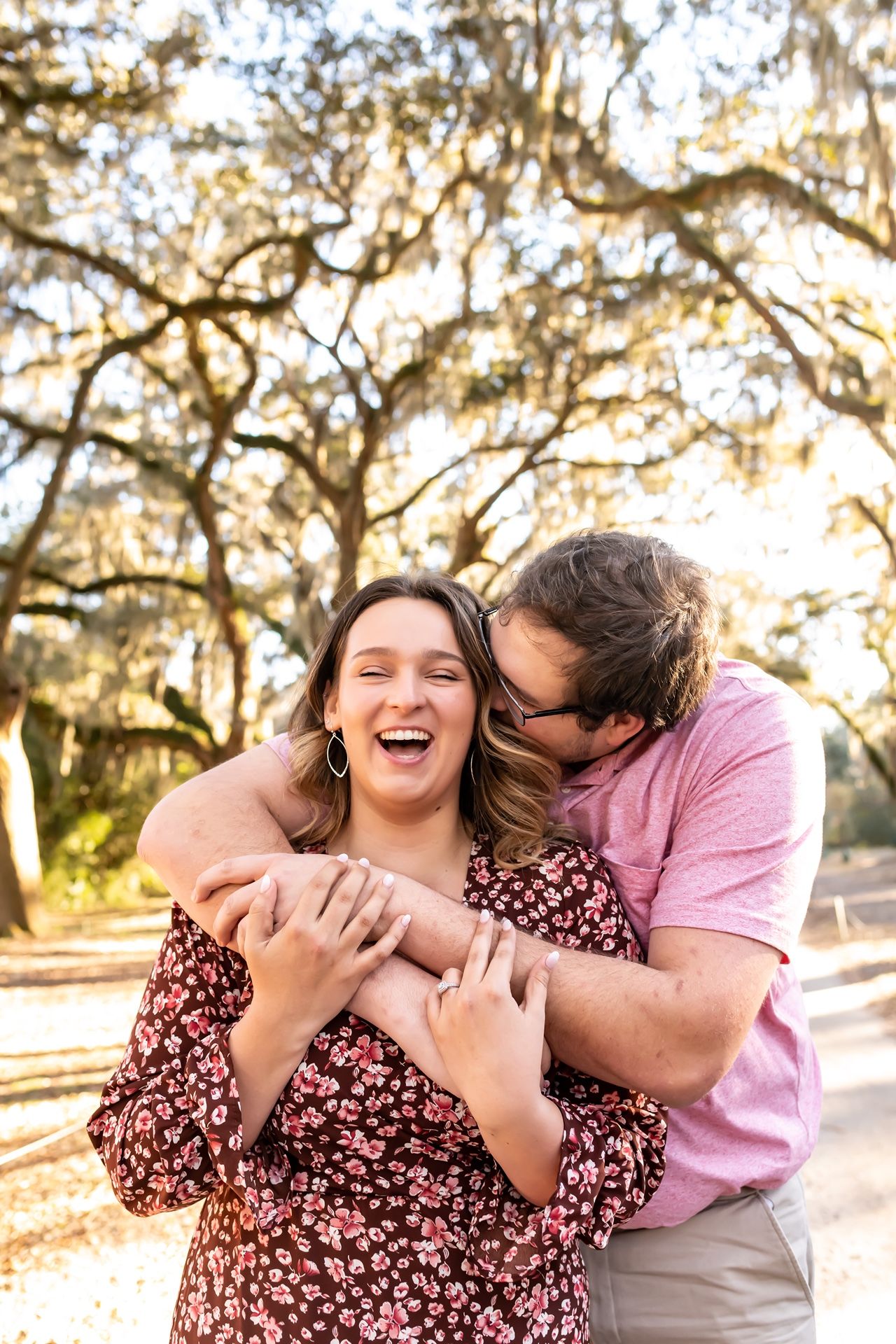 A couple shares a warm embrace and joyful laughter against a backdrop of sunlit trees with dangling Spanish moss.