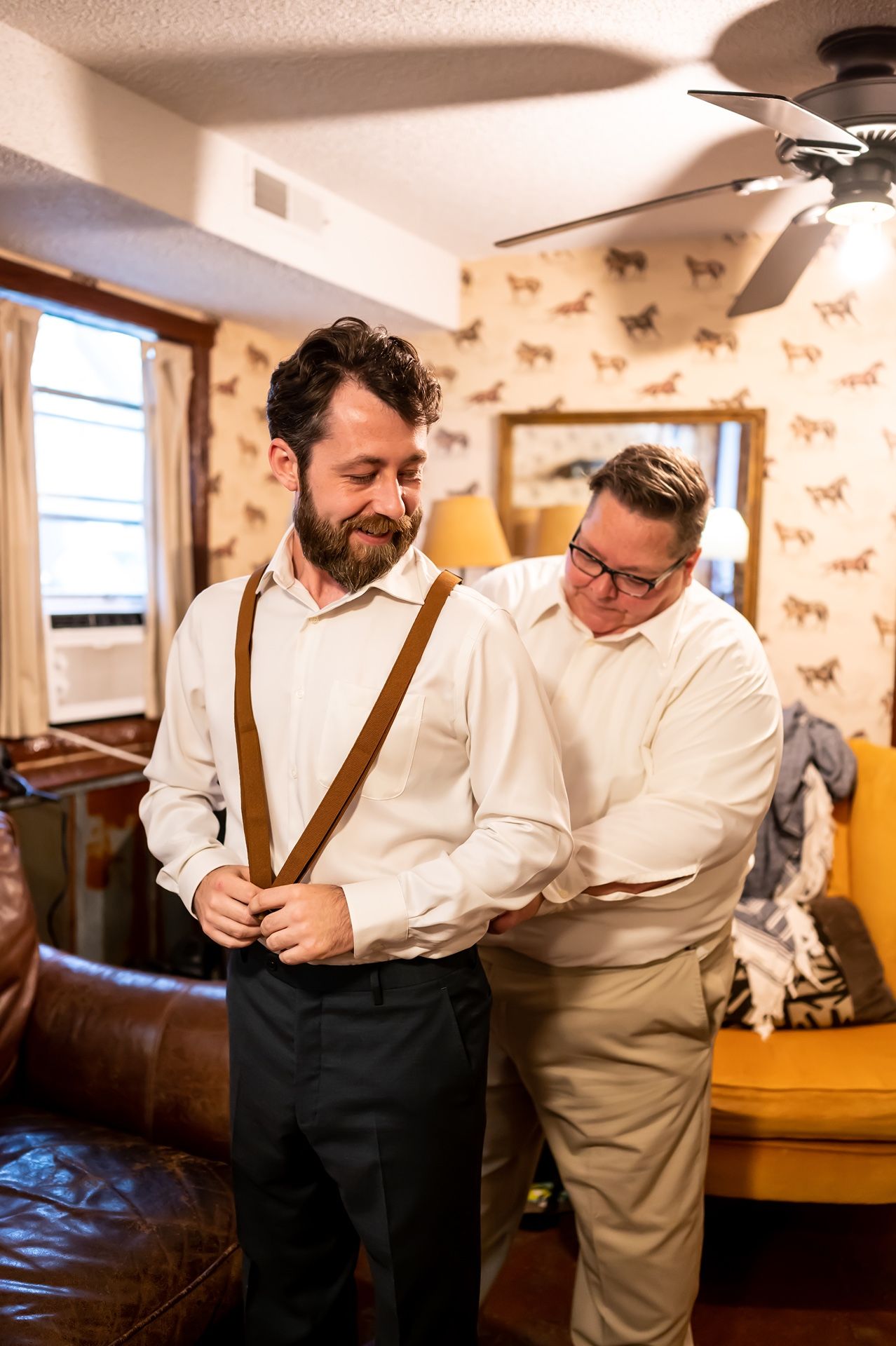 Two people in a room with horse-print wallpaper help with formal attire preparations and adjusting brown suspenders.