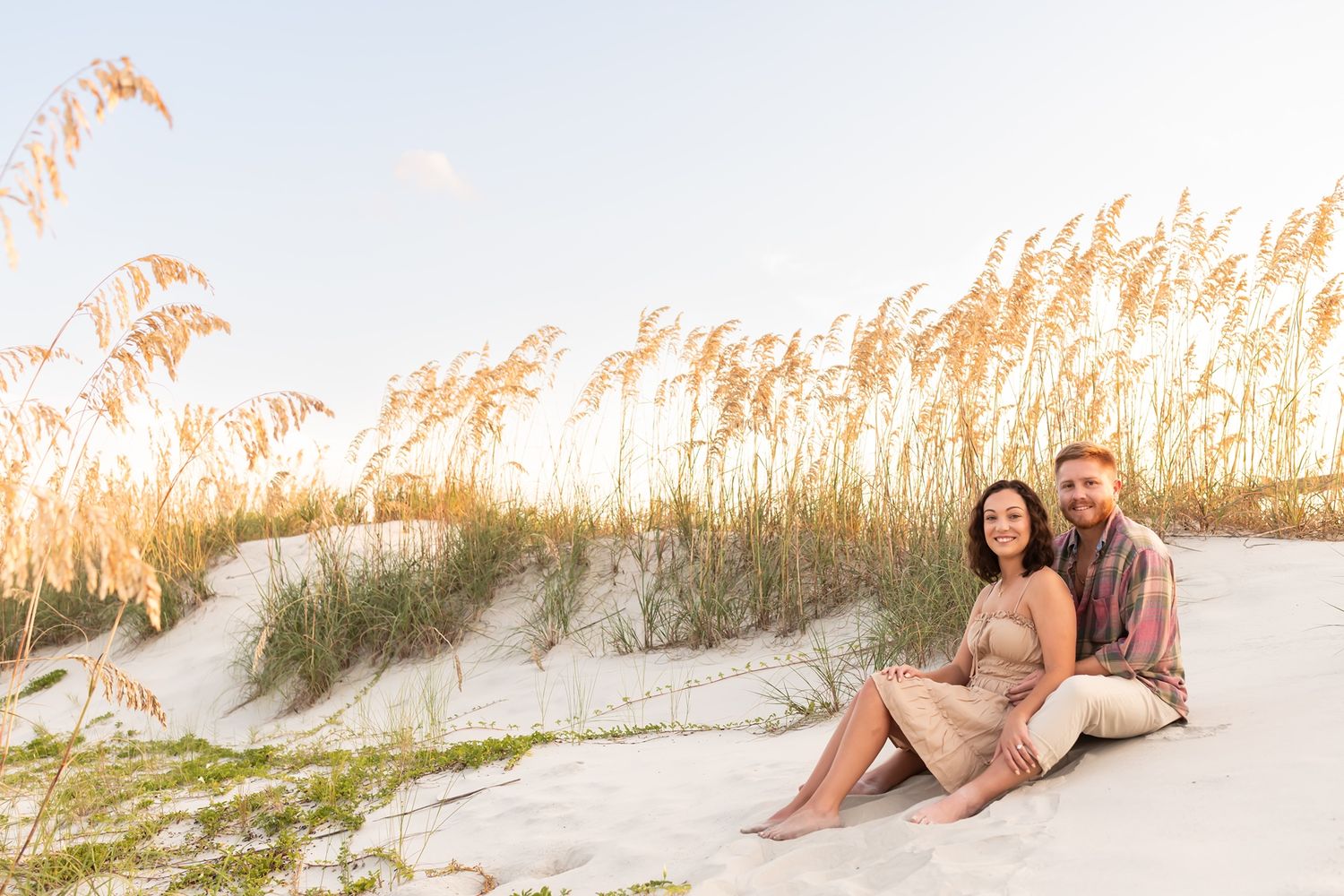 A couple sits together on white sand dunes at sunset with golden sea oats swaying in the background.