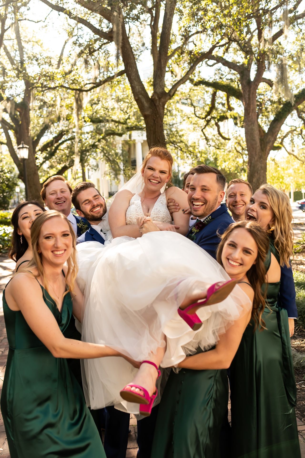 A playful wedding party lifts the bride wearing bright red shoes under spanish moss draped trees.