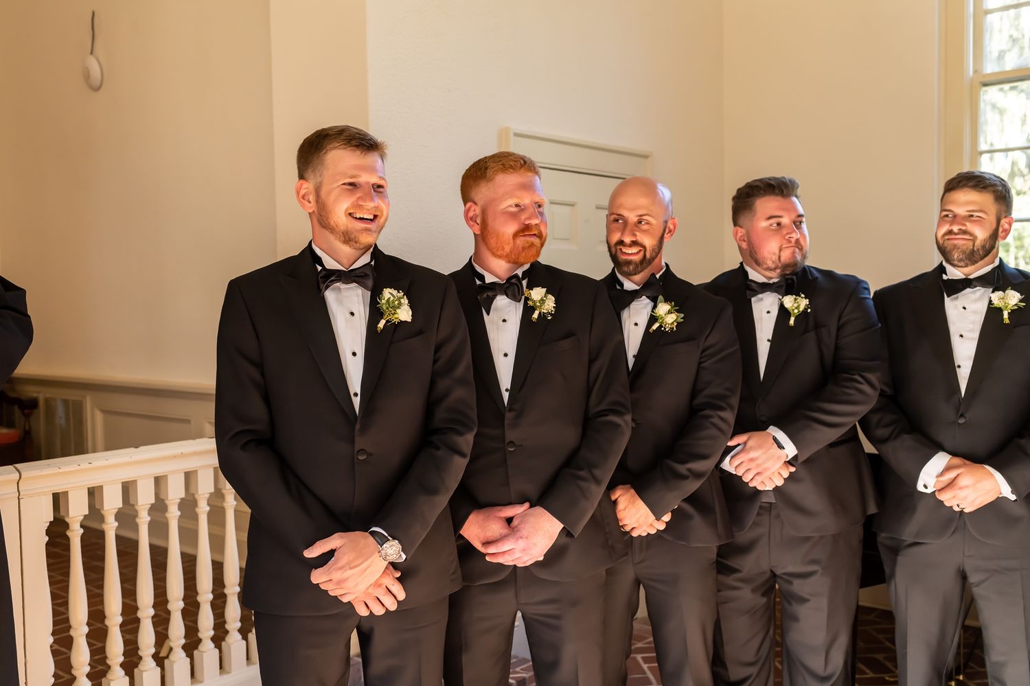 A row of groomsmen laughing while standing together in formal black tuxedos at a wedding venue.