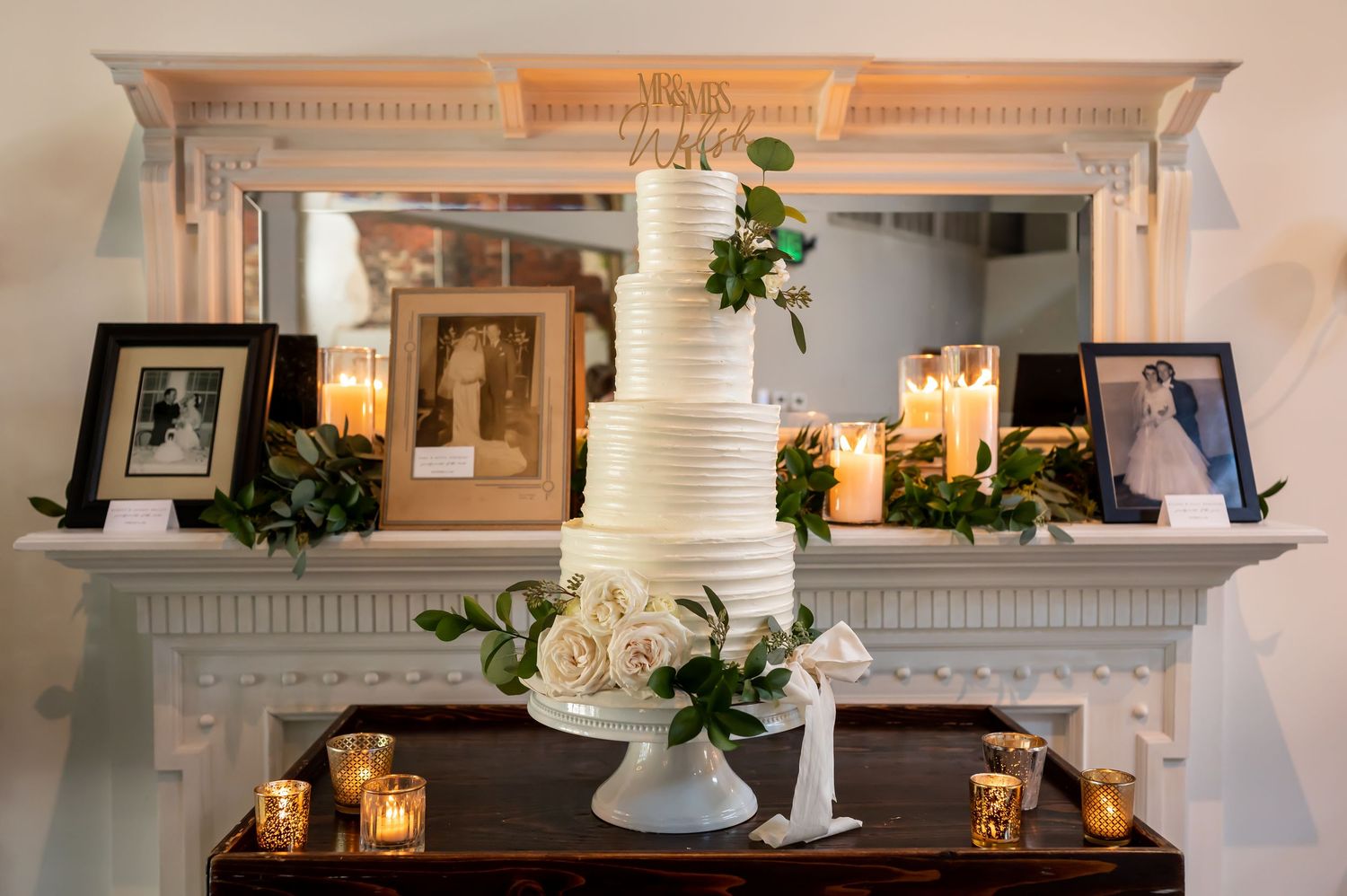 Elegant white wedding cake displayed on ornate mantel with framed photos, candles and floral decorations.