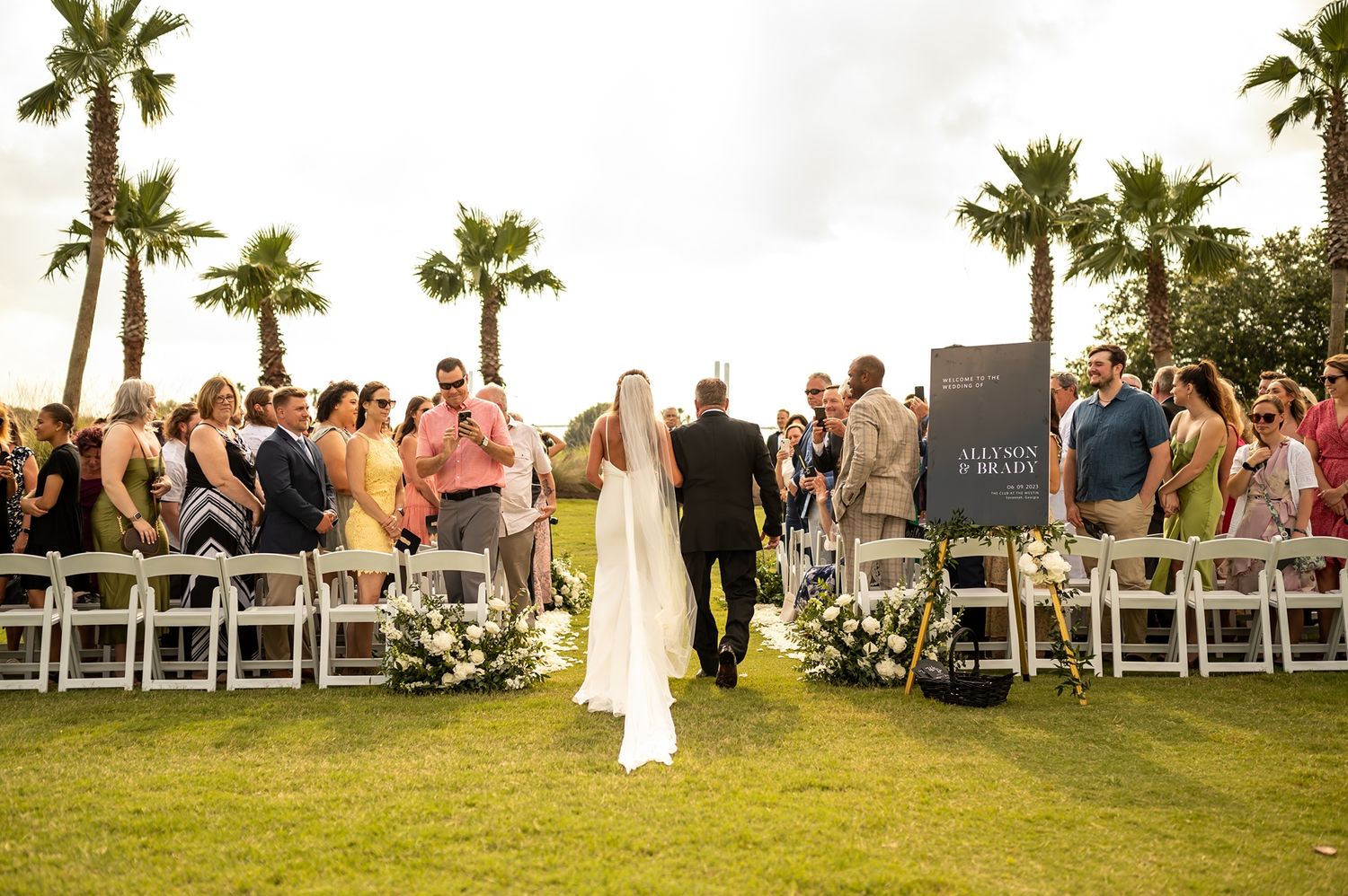 Wedding ceremony on a lush green lawn with palm trees as guests gather to witness the romantic outdoor occasion.