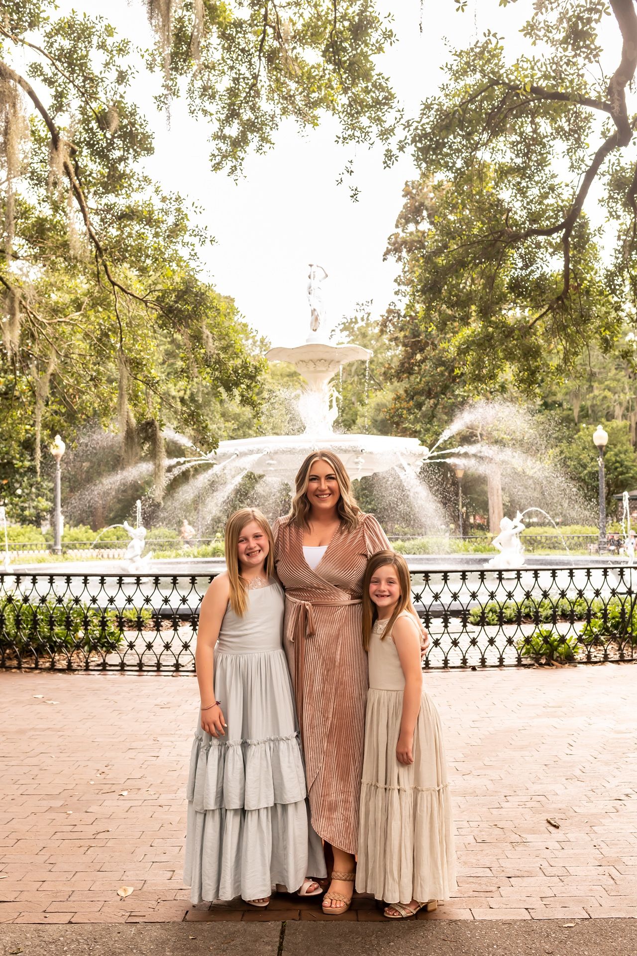 Three people stand in front of a beautiful fountain in a historic park with Spanish moss hanging from trees.