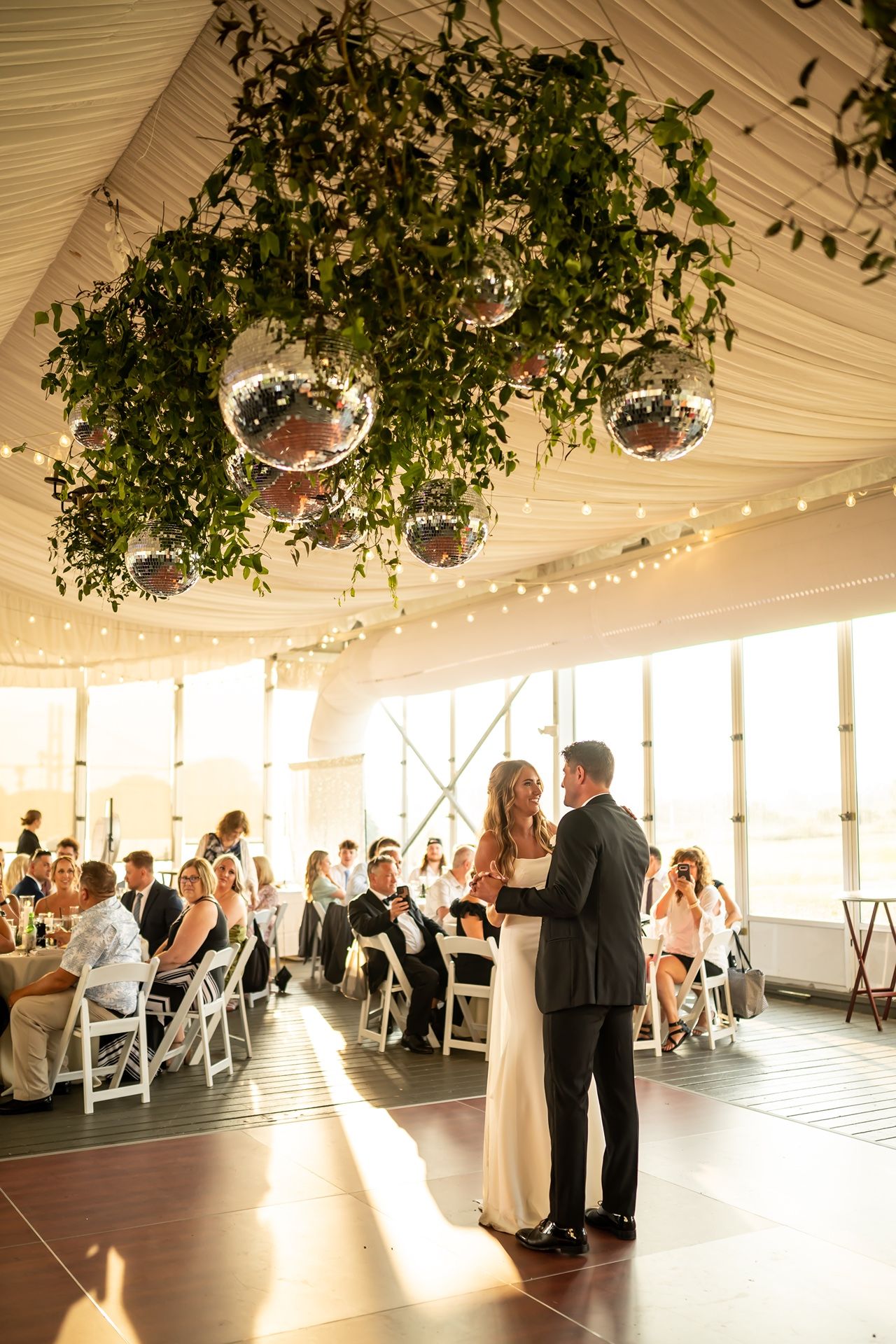 A wedding couple shares their first dance under disco balls and greenery in a bright sunlit reception venue.
