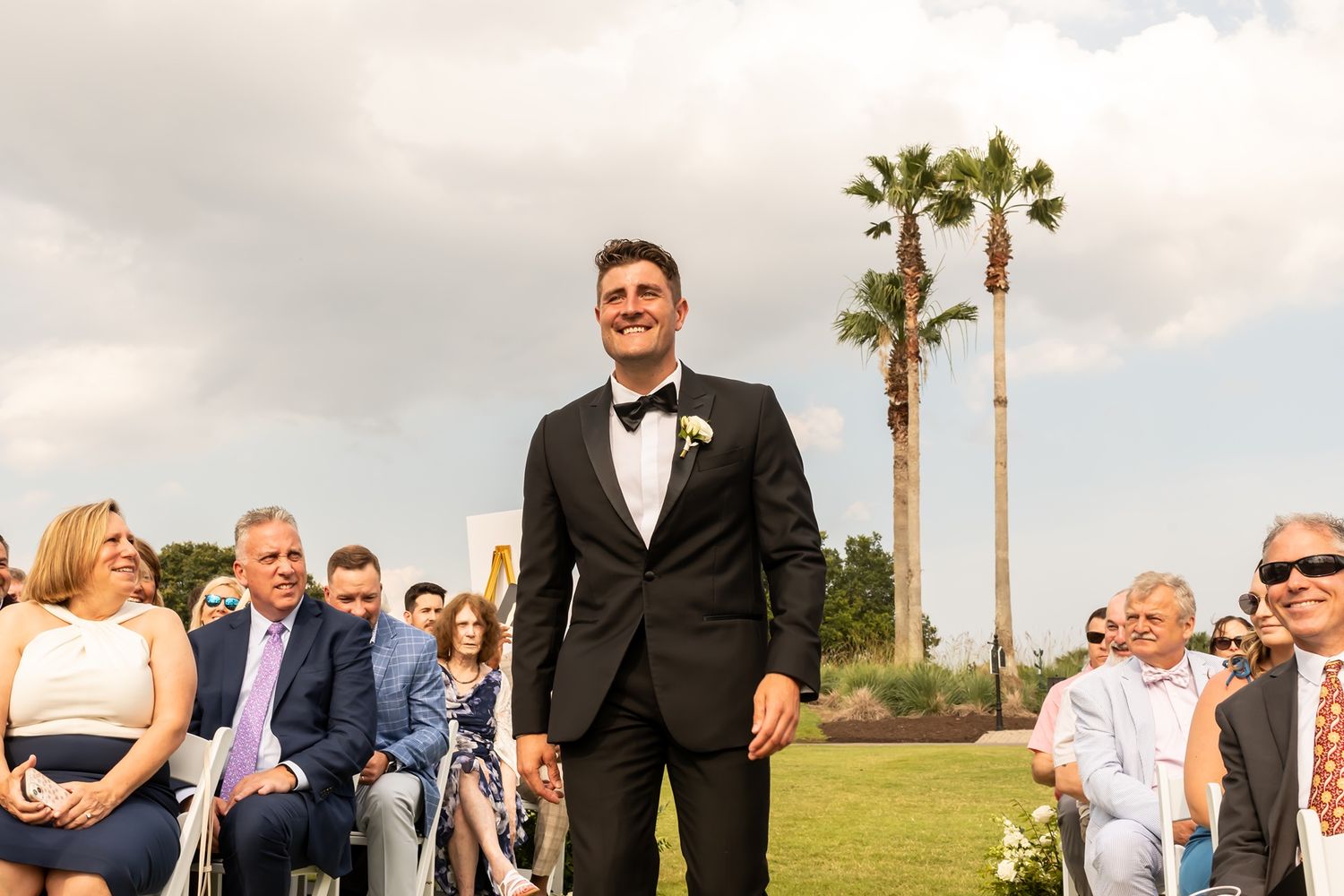 Cheerful groom stands at outdoor wedding ceremony with palm trees and seated guests in background.