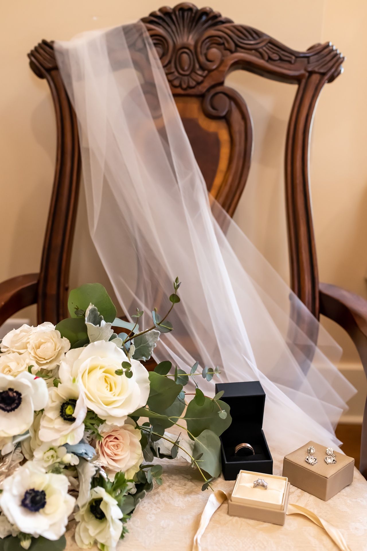 Wedding details including floral bouquet with white roses, sheer veil, and ring boxes displayed on antique wood chair.