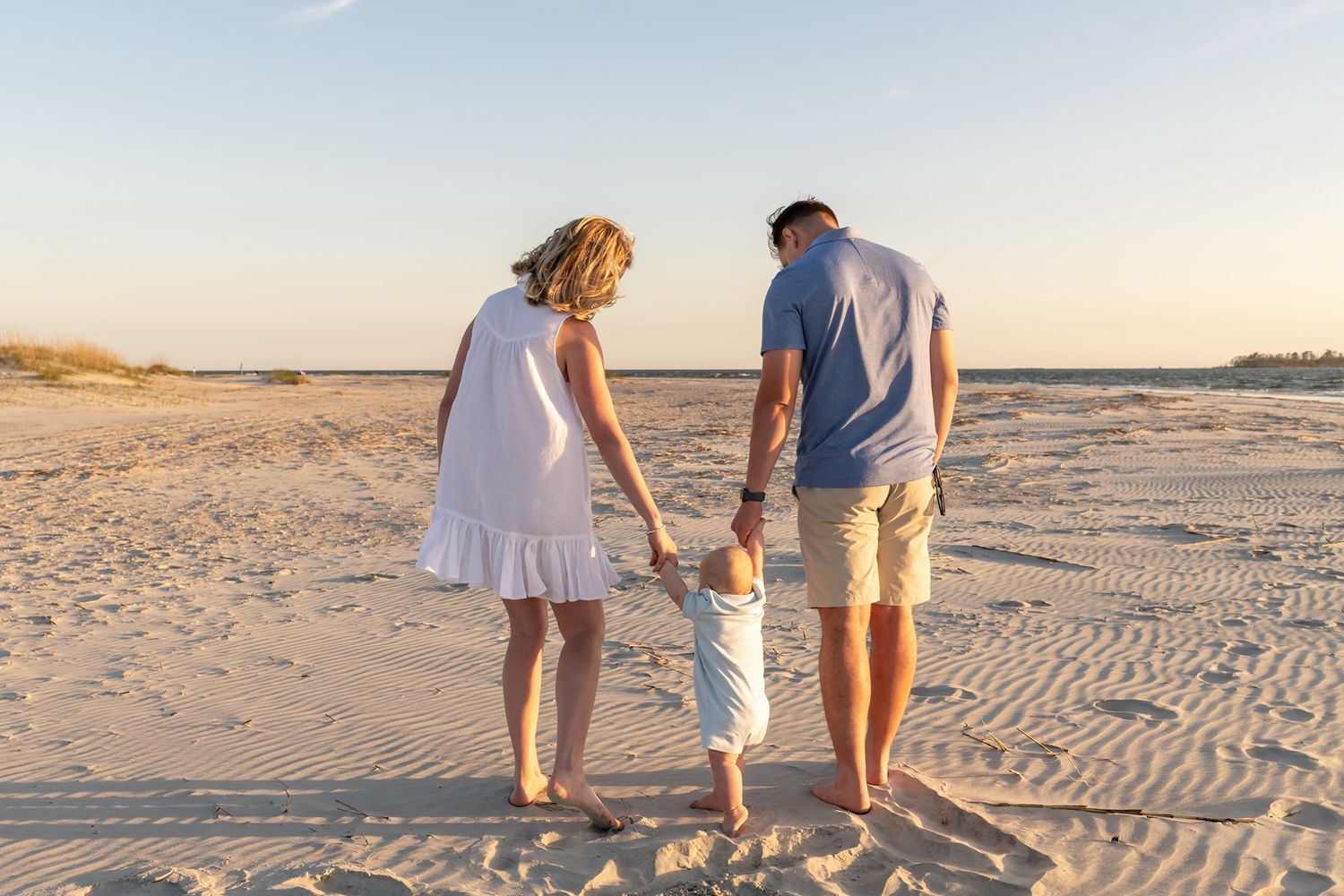 Three people walk together on a sandy beach at sunset, holding hands while leaving footprints in the sand.
