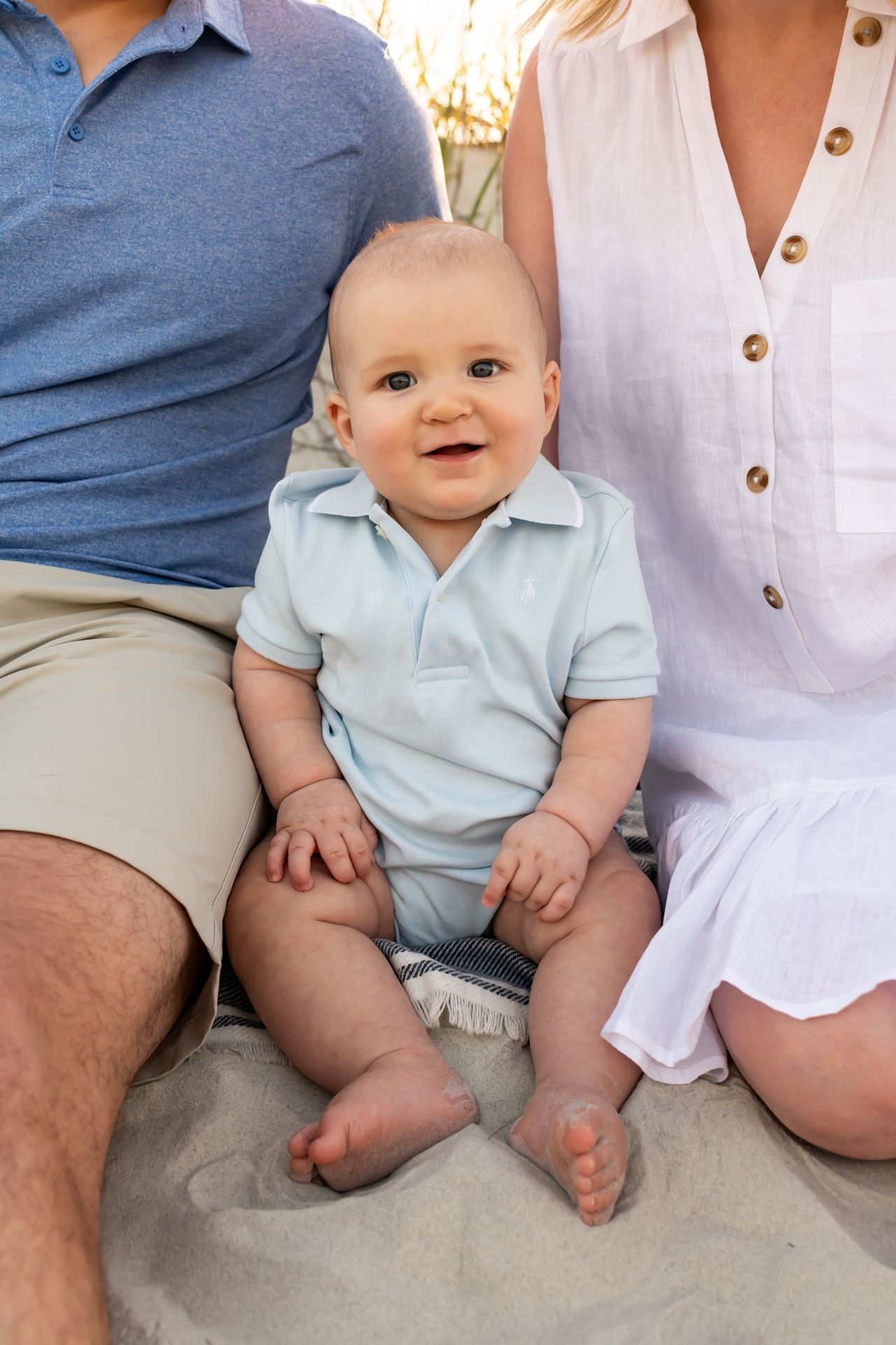 Joyful baby in light blue polo sitting on sand between their seated companions outdoors at sunset.