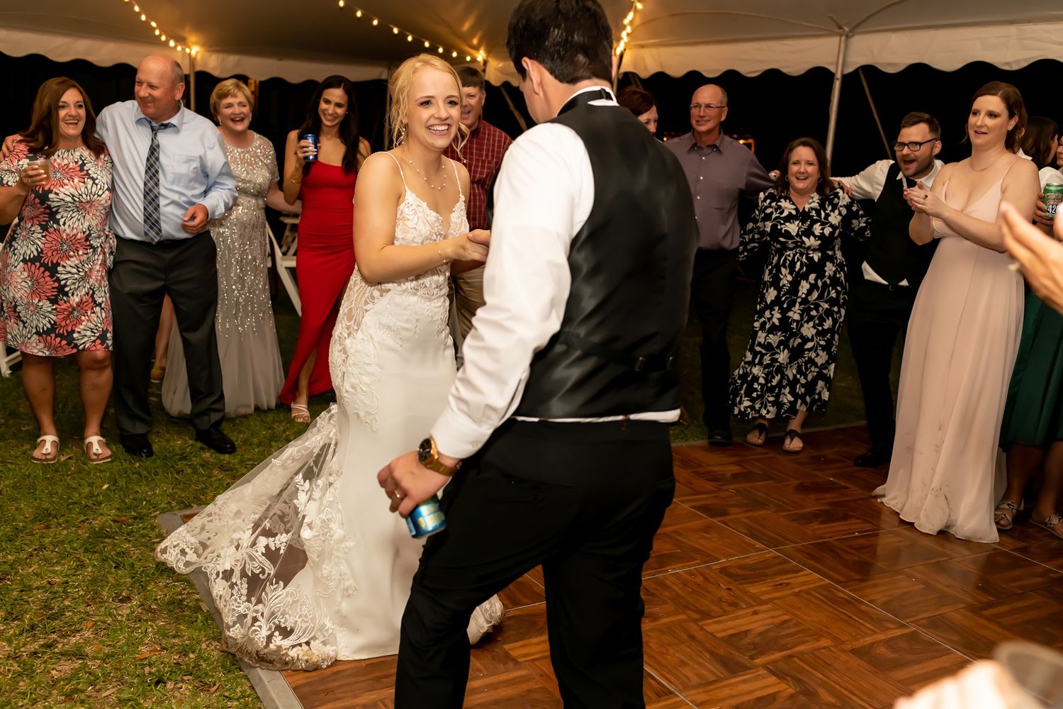 A couple dances together on a wooden dance floor under a tent while wedding guests watch and smile at the celebration.