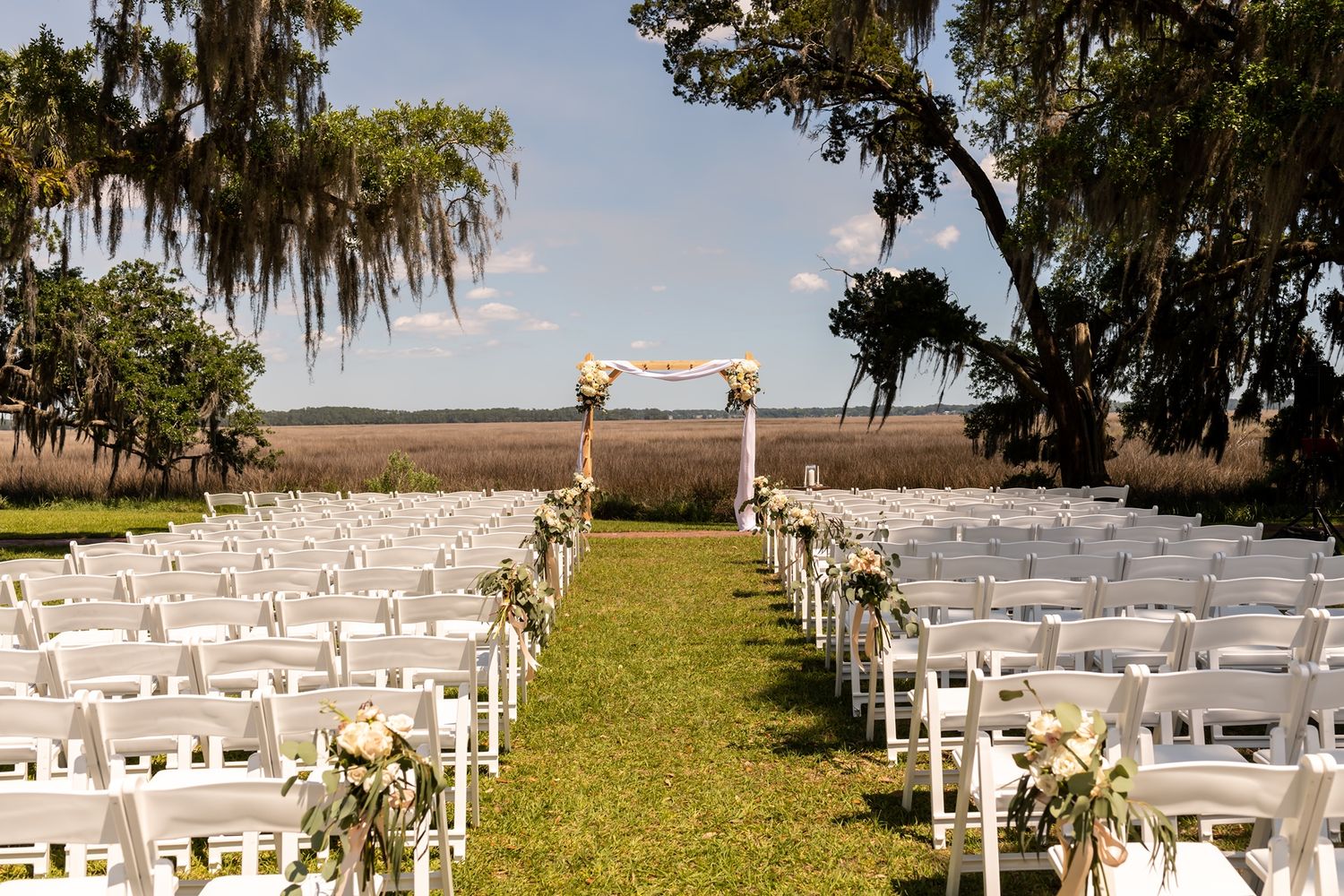 An outdoor wedding ceremony setup with rows of white chairs and Spanish moss draped trees framing a waterfront view.