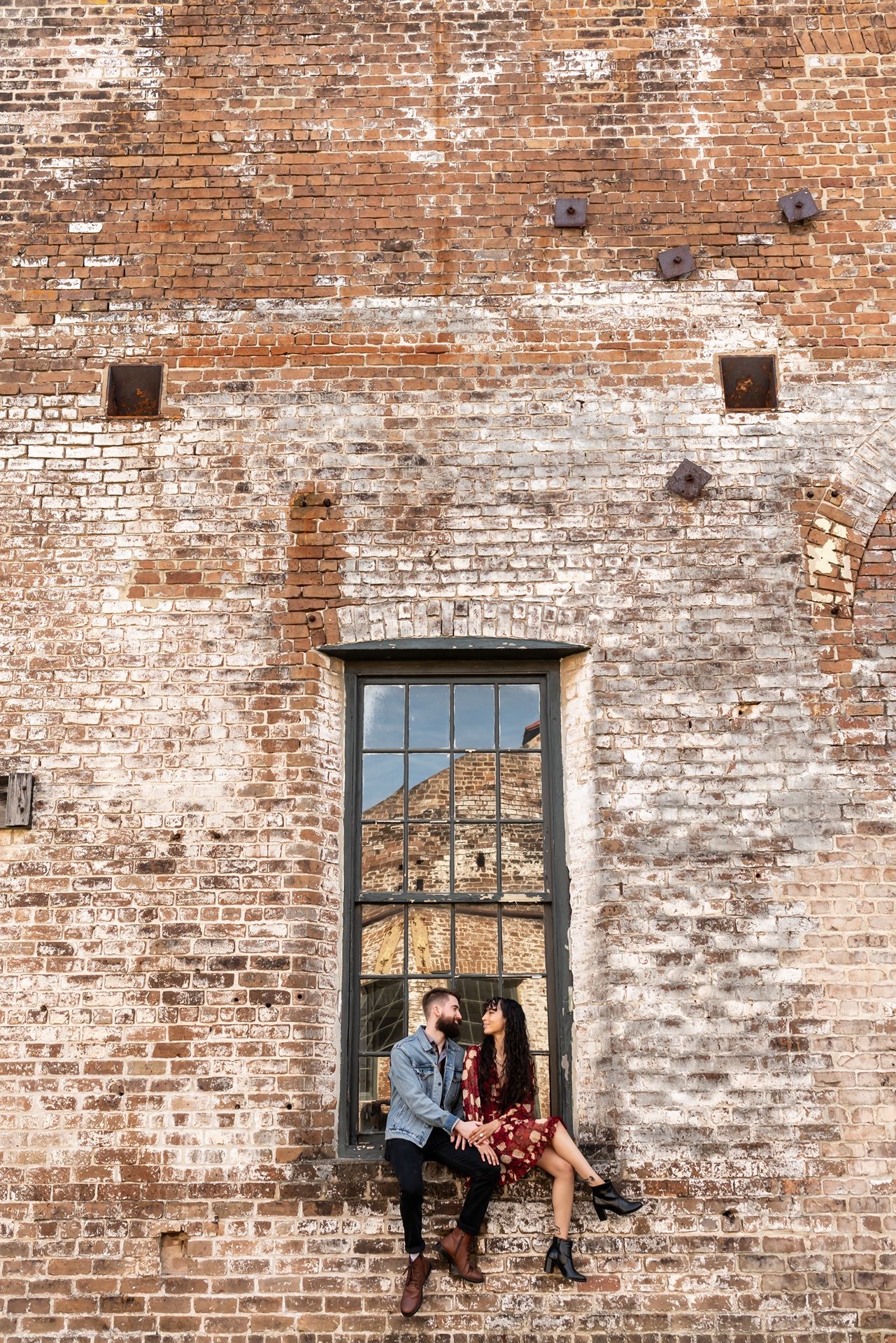 Couple sits in a rustic window frame of an old brick building with weathered textures and industrial charm.