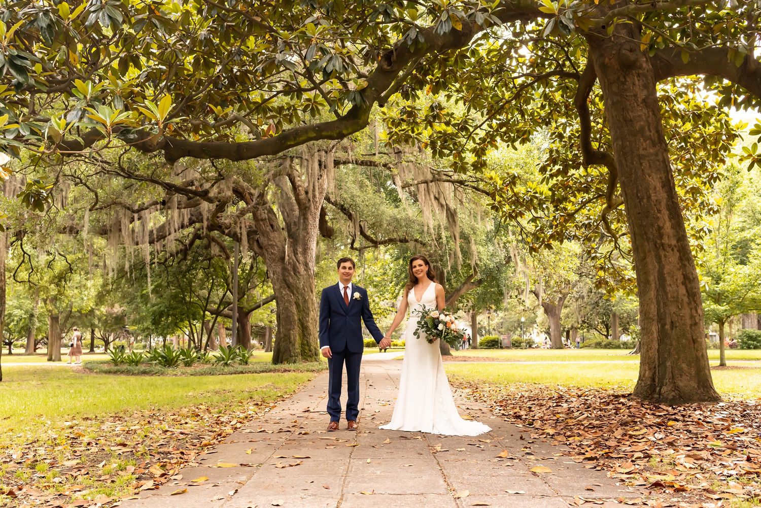 A couple walks together along a tree-lined path scattered with autumn leaves in a peaceful garden setting.