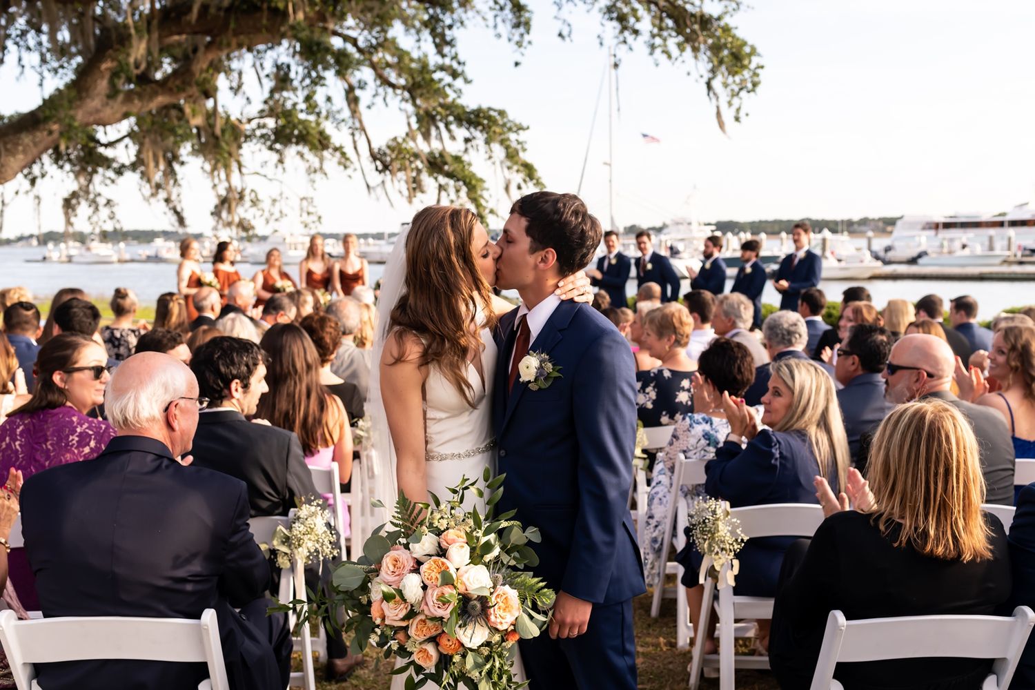 Wedding guests seated in white chairs at an outdoor ceremony overlooking a waterfront view.