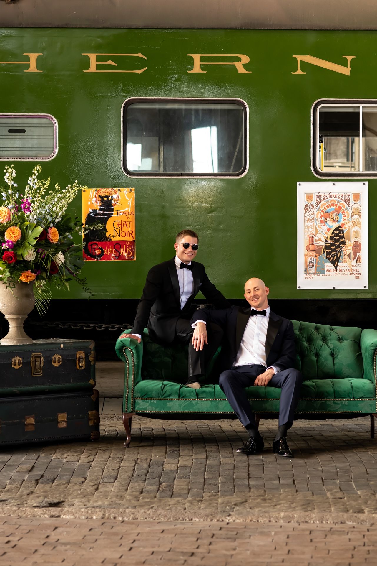 Two people pose on a green velvet couch in front of a vintage green train car with retro posters.