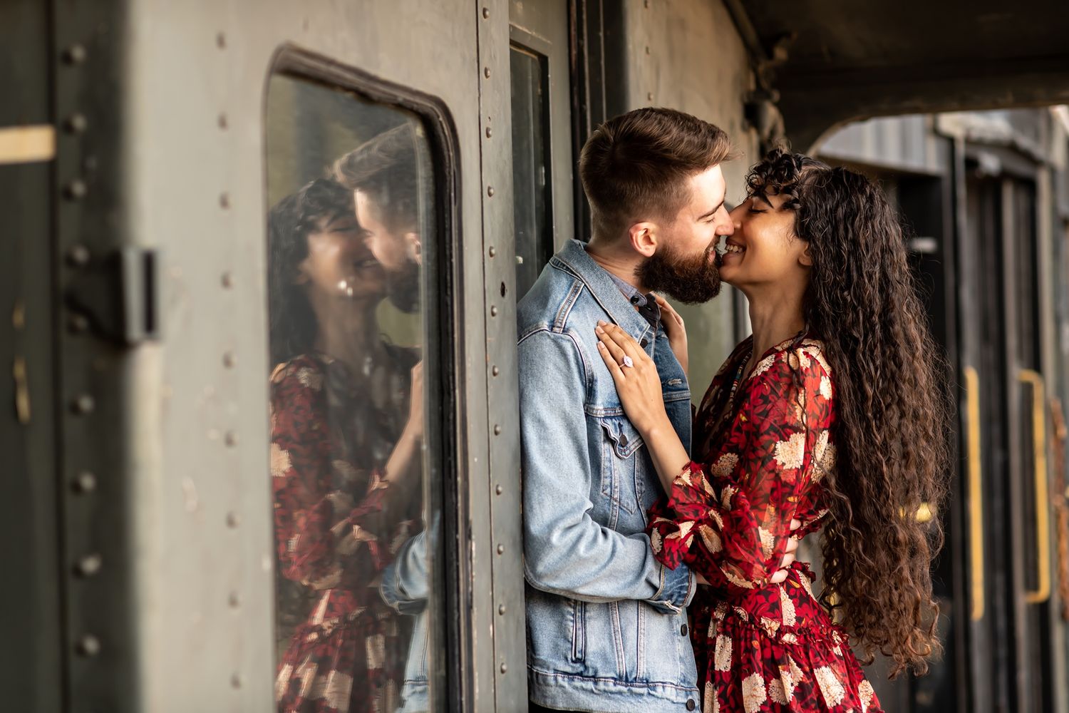 Romantic couple sharing an intimate moment by a vintage train car door wearing denim and red floral dress.