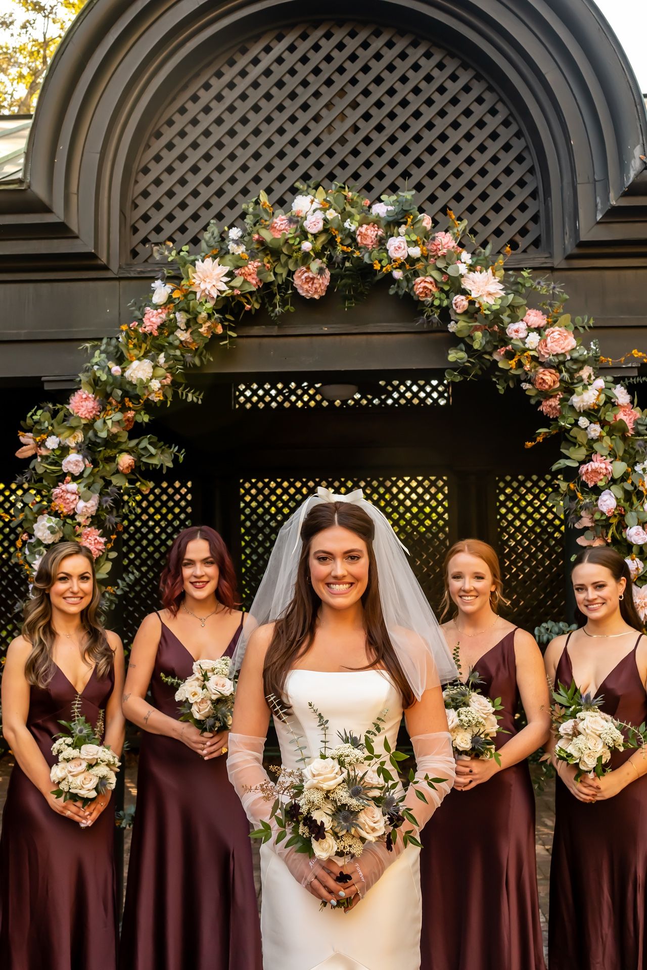 A wedding party poses outside an elegant gazebo adorned with pink and white floral arrangements against a lattice backdrop.
