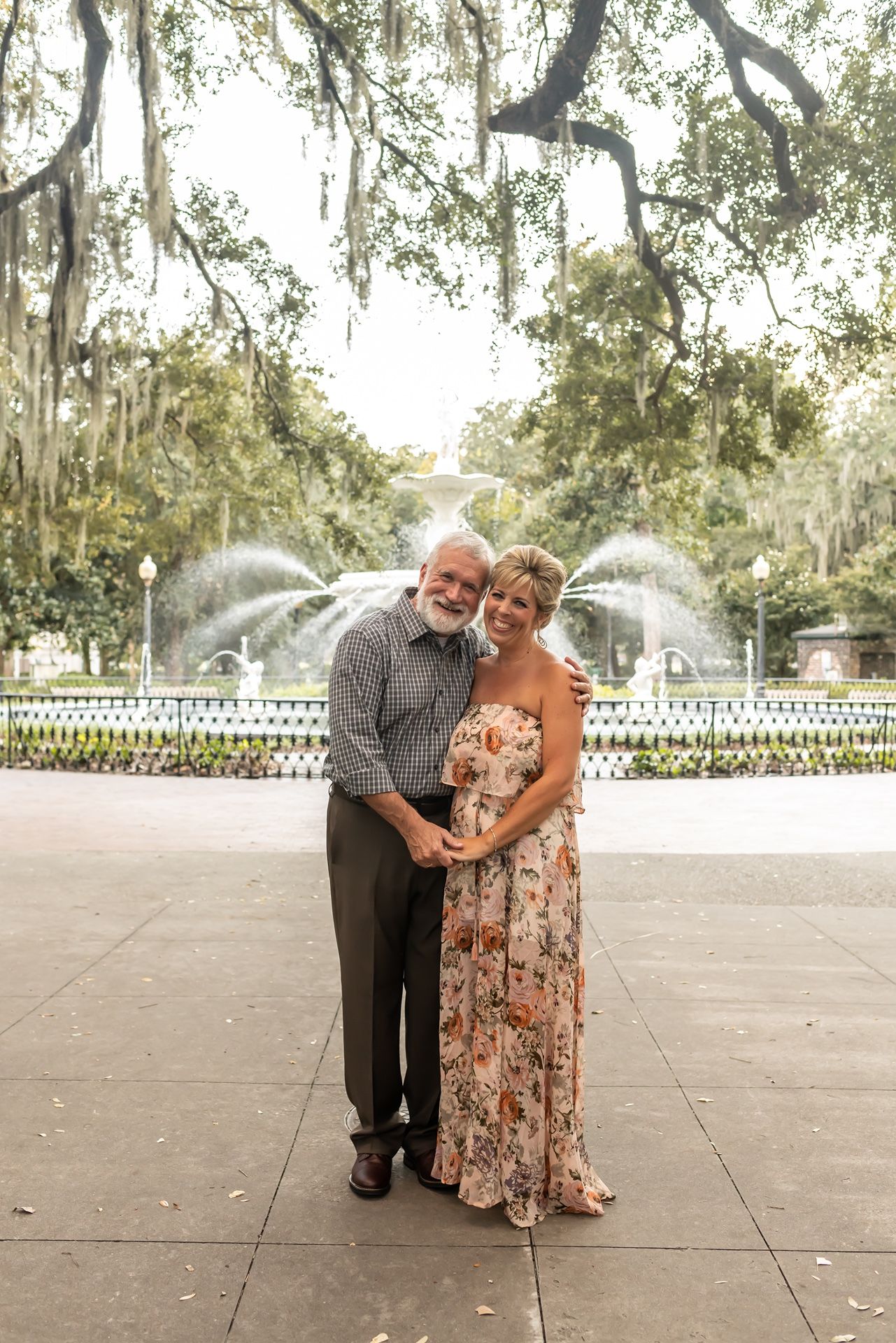 A couple poses together in front of a beautiful fountain in a lush garden setting.