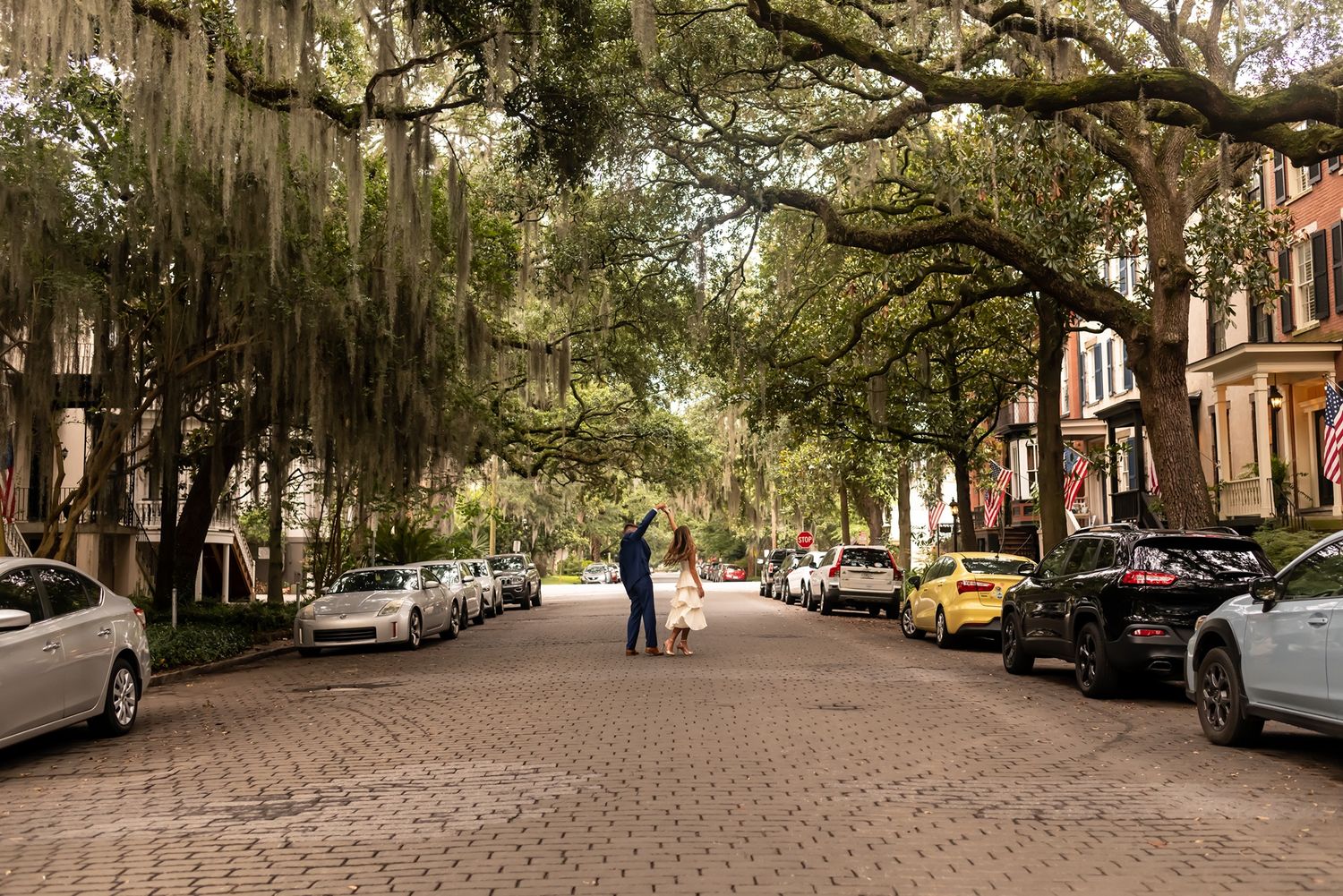 Historic street in Savannah lined with Spanish moss-draped oak trees, parked cars and brick paving stones.