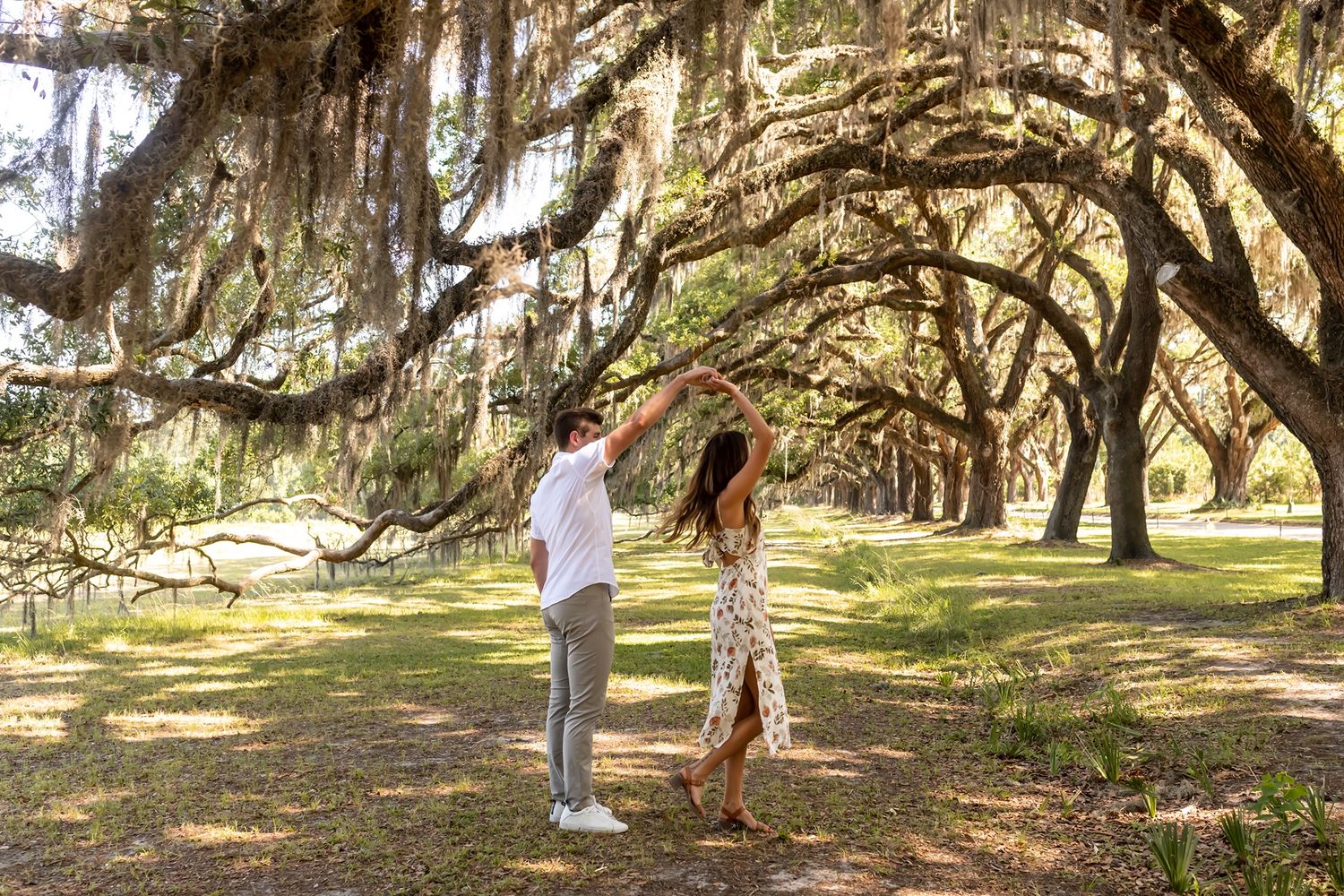 Two people stand beneath majestic live oak trees draped with spanish moss in a scenic southern garden setting.