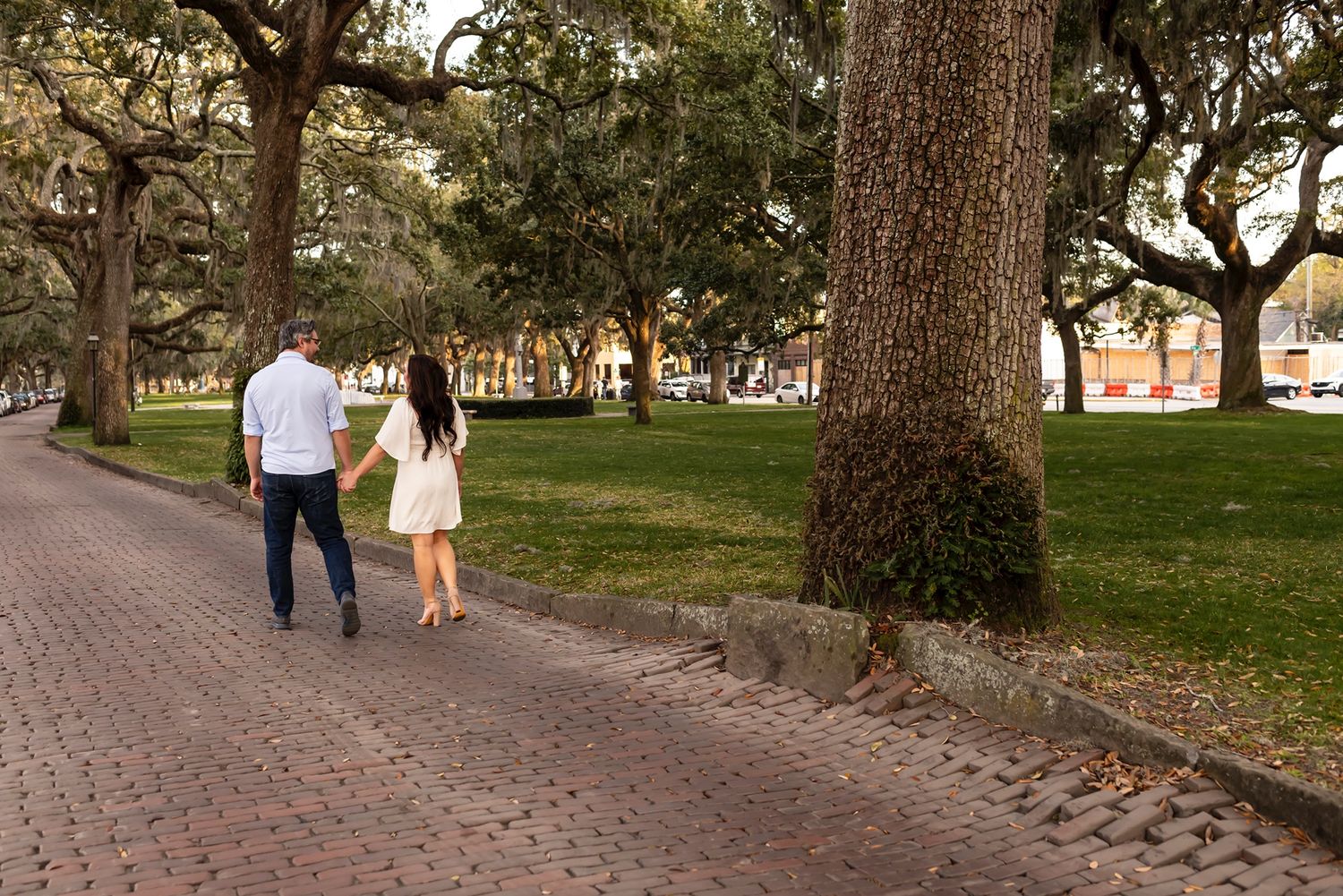A couple walks hand in hand along a cobblestone path under large oak trees in a peaceful park setting.