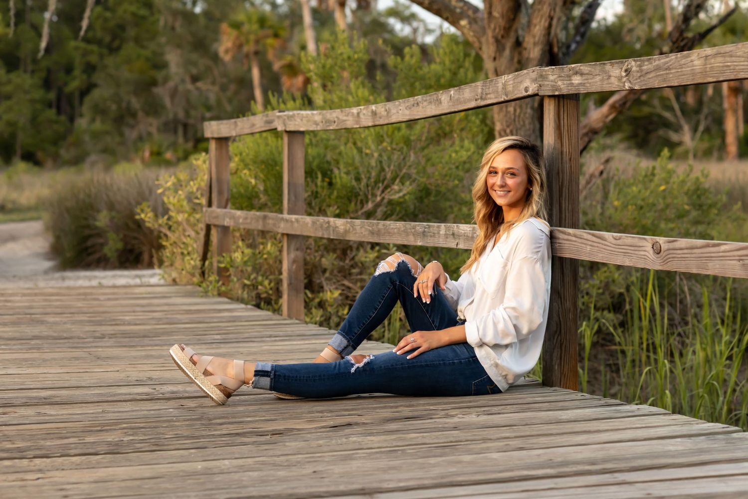 Someone sitting casually on a wooden boardwalk by marsh grasses on a sunny afternoon wearing jeans and a white shirt.