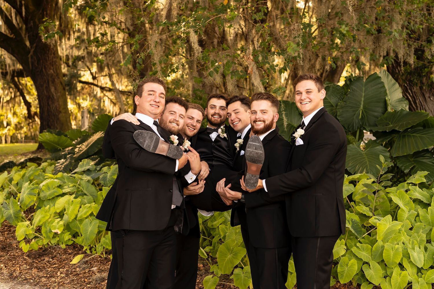 A playful groomsmen photo showing men in black tuxedos posing with lifted arms in an outdoor garden setting.