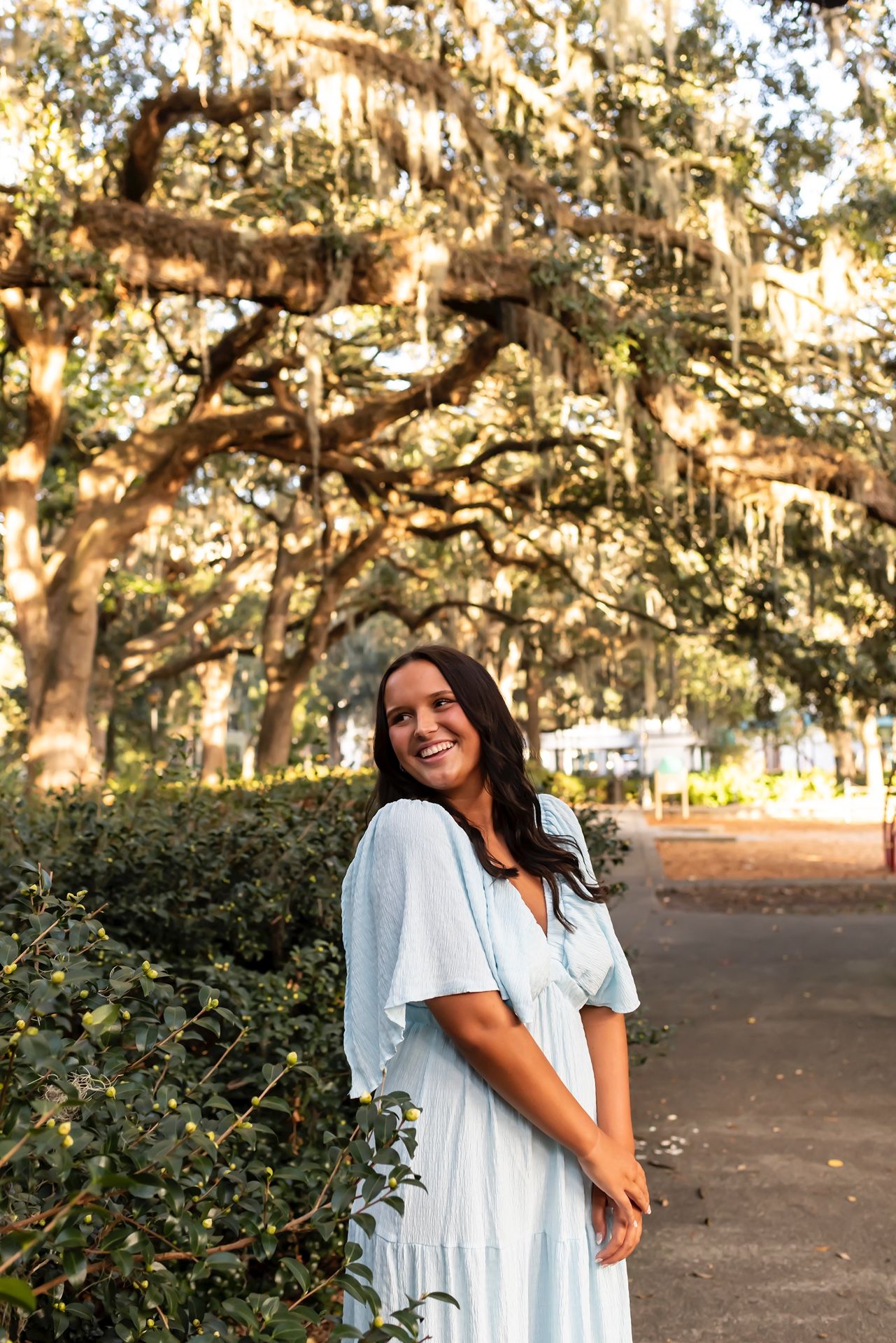 Woman in white flowy dress poses outdoors among Spanish moss-draped trees during golden hour in a park setting.