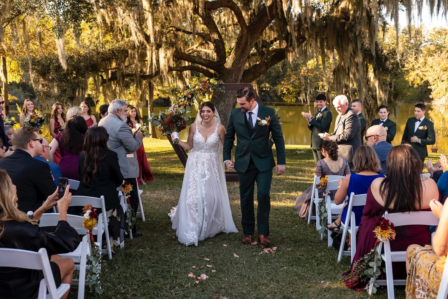 An outdoor wedding ceremony under oak trees with Spanish moss as newlyweds stand before seated guests.