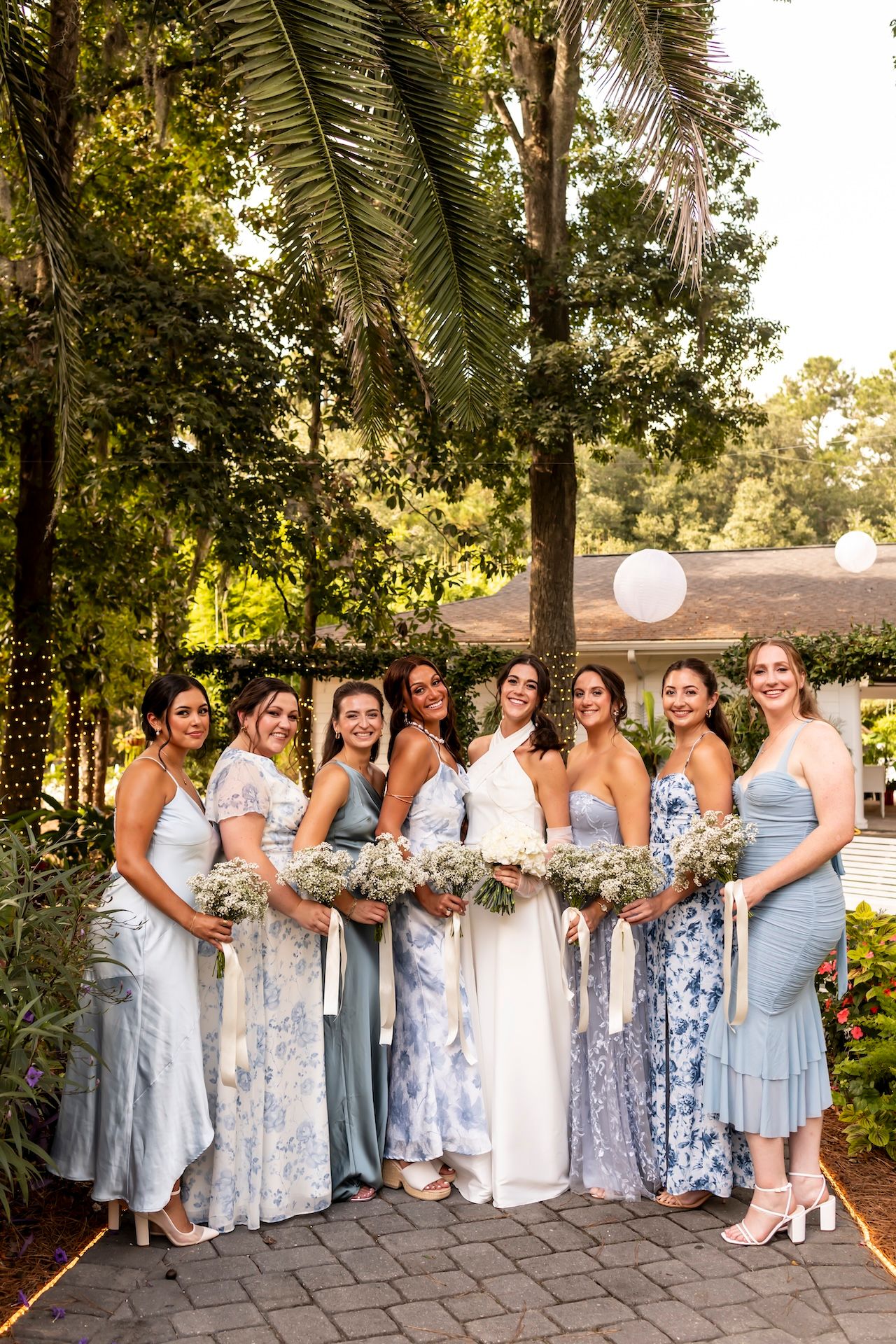 A bride in white stands with seven bridesmaids in blue dresses holding bouquets outdoors among lush green trees.