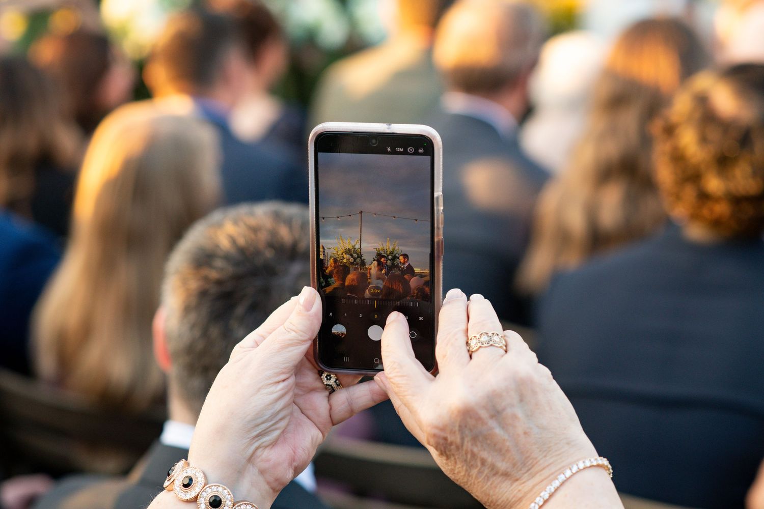 Hands holding smartphone to take photo of crowd at outdoor event during golden hour lighting.