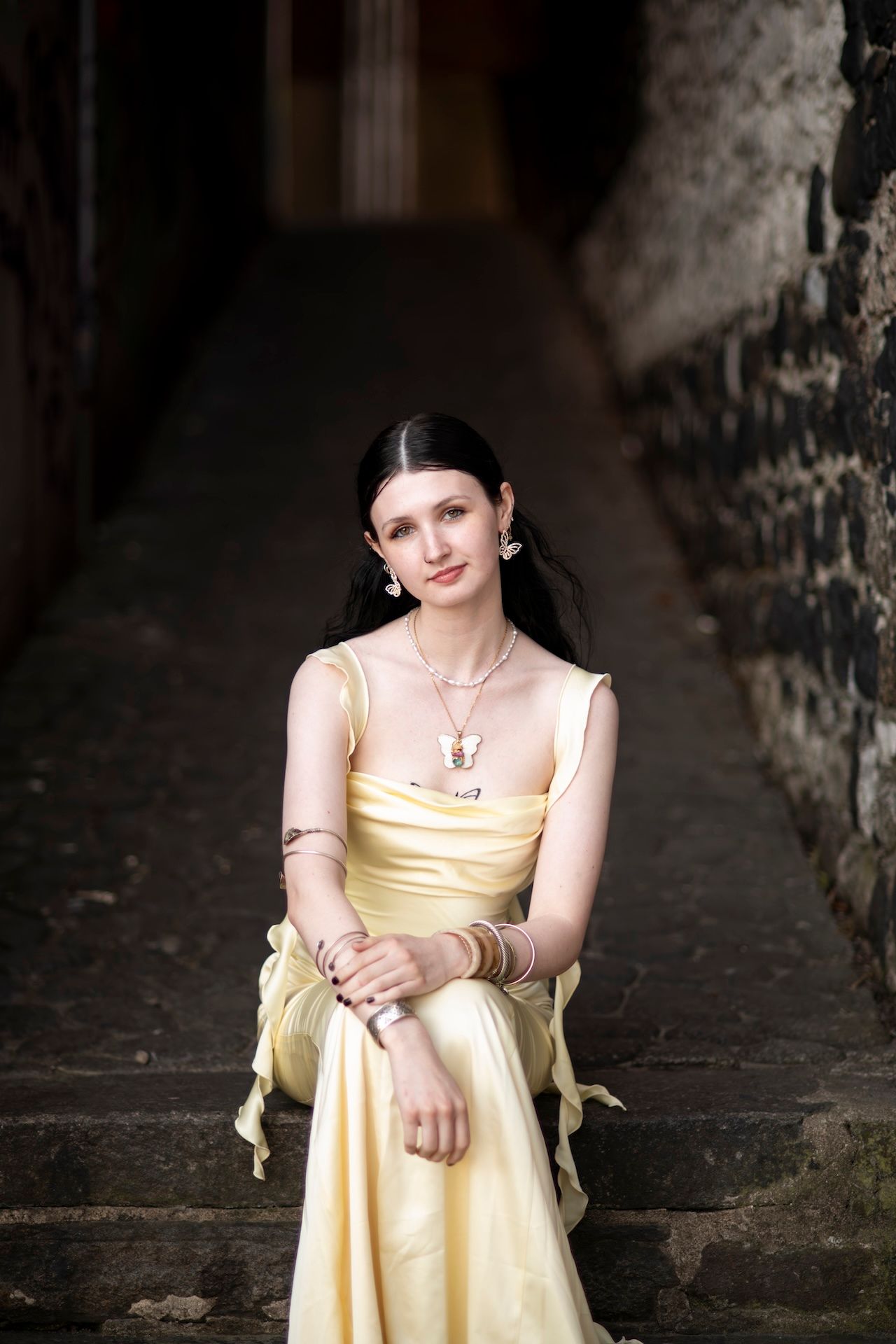 Young woman in yellow satin dress sitting on stone steps, wearing gold jewelry and bracelets.