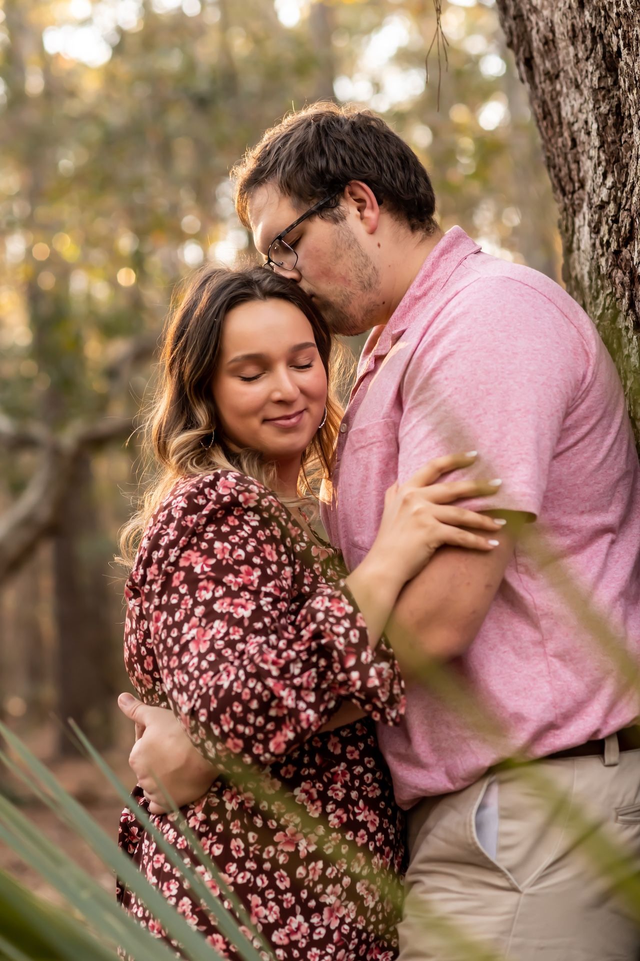 A romantic couple embraces outdoors in a wooded setting, wearing pink and floral clothing during golden hour.