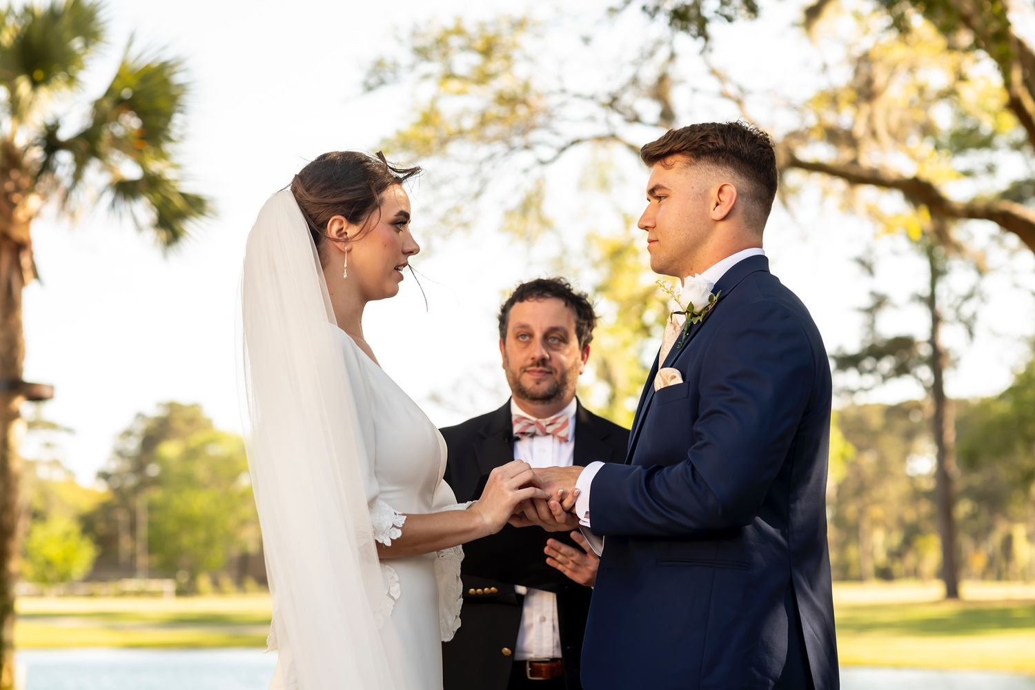 Wedding ceremony participants exchange rings in an intimate garden setting with natural lighting.