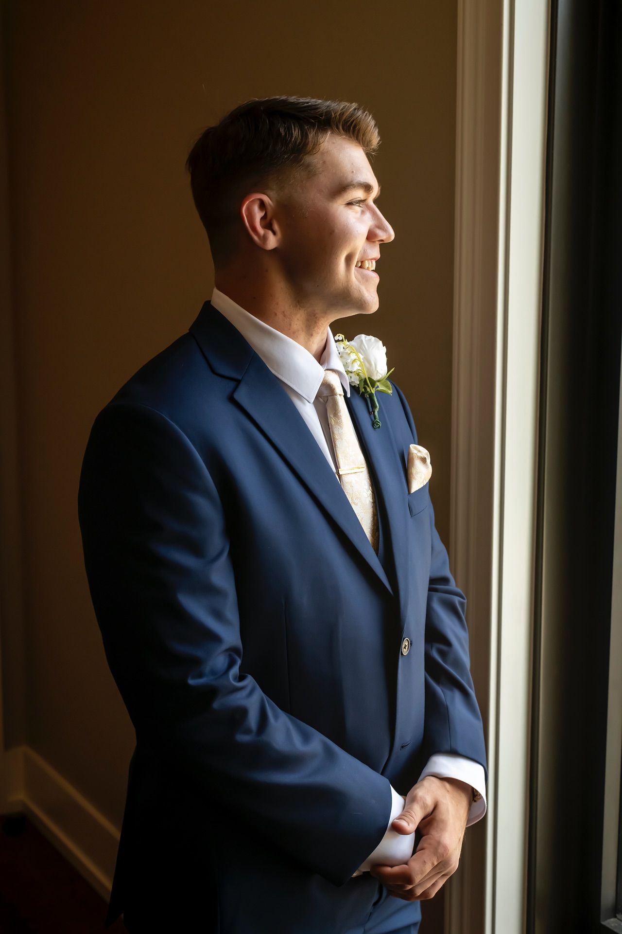 Side profile of someone in a navy blue suit with boutonniere looking thoughtfully through window in natural light.